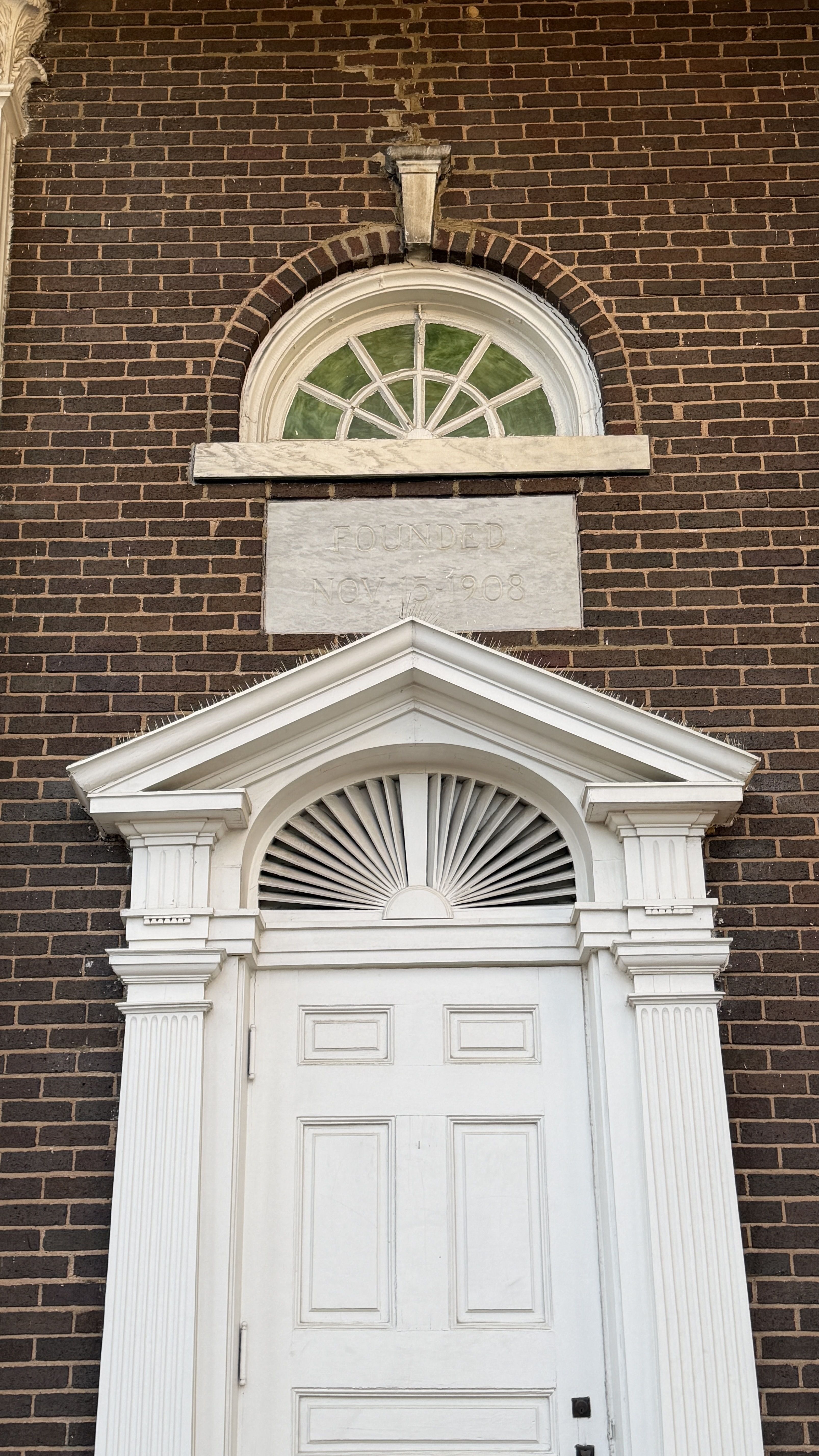 White wooden door with detailed trim and a fanlight window above, set in a red brick wall. A plaque above the door reads "FOUNDED NOV 15 1908".