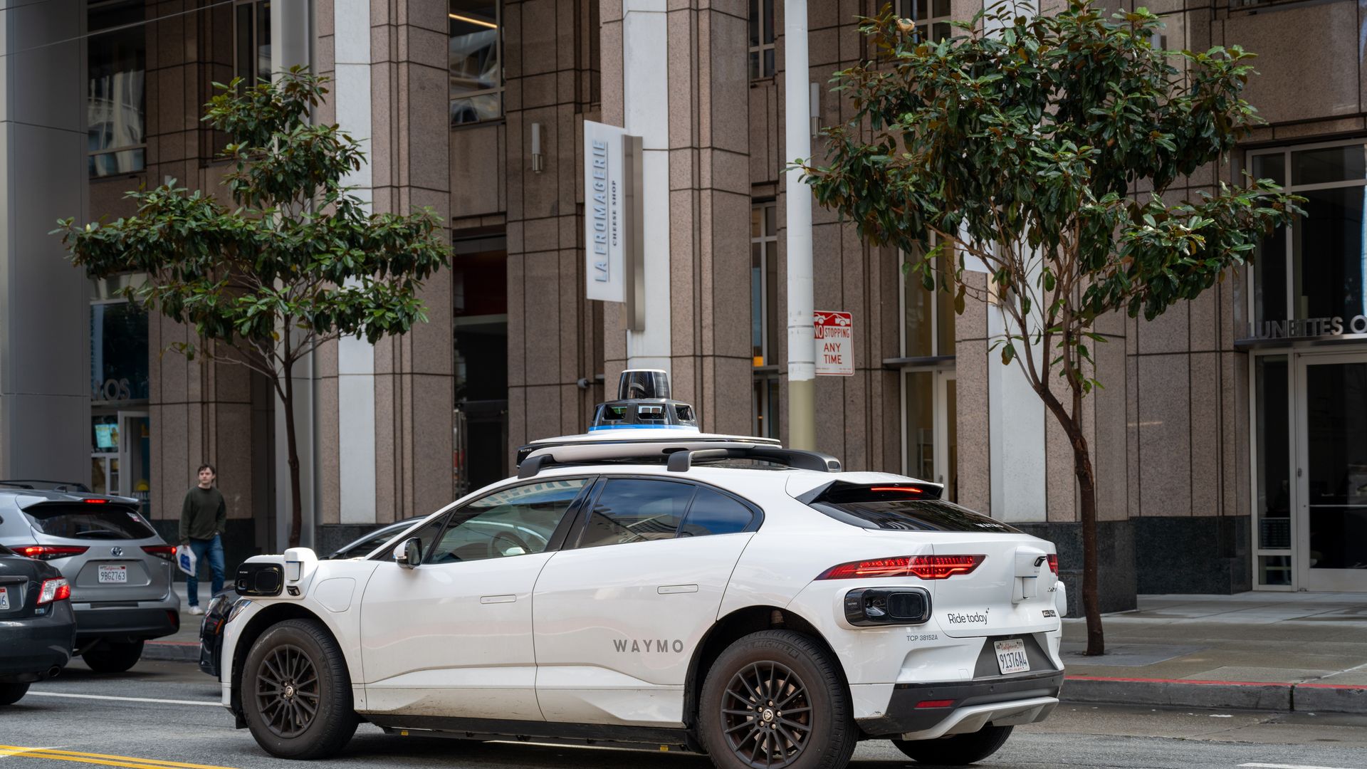 White Waymo autonomous electric vehicle equipped with multiple sensors on roof and sides parked on city street near trees and office buildings.