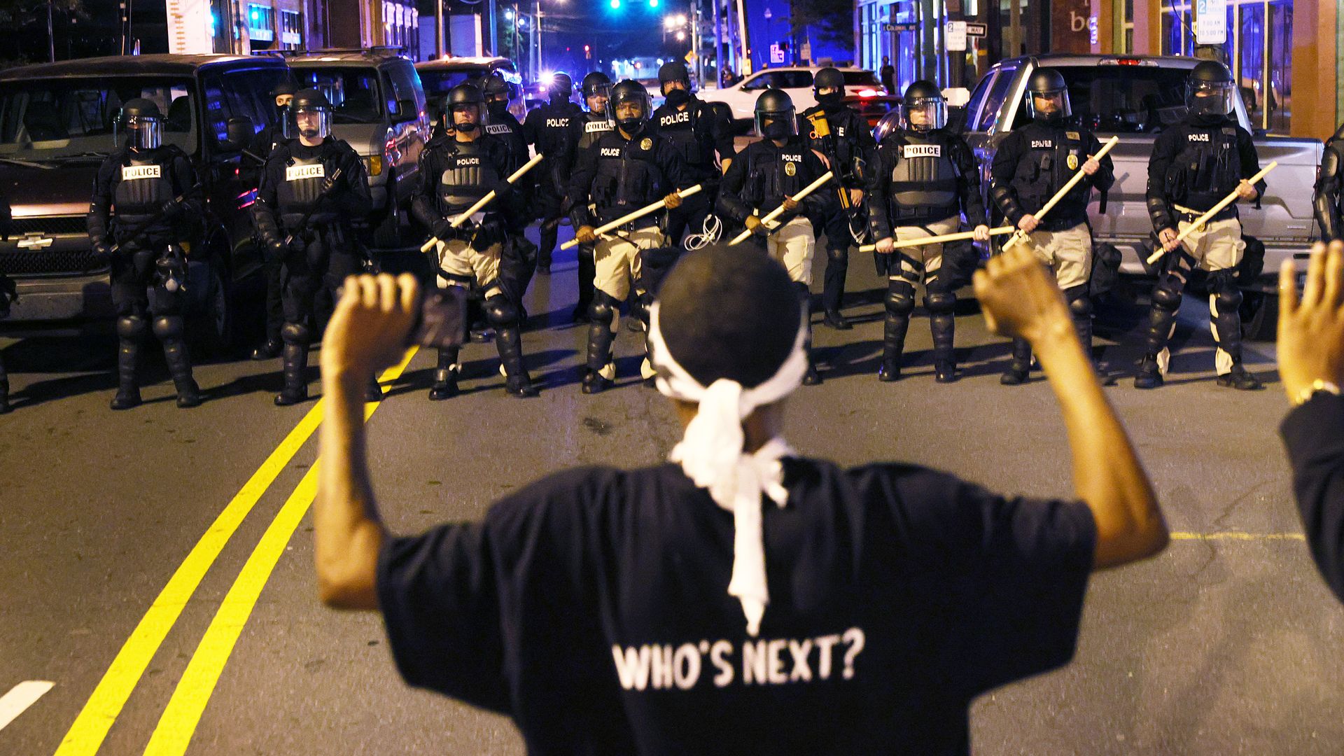 Photo of a protester wearing a shirt that says "Who's next" as they face off against police in riot gear