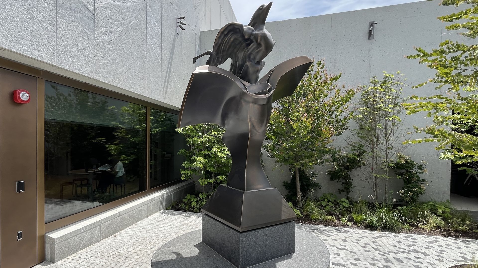 Metal sculpture of a bird with wings spread, perched on an open book, mounted on a circular pedestal outside a modern gray building with glass windows and greenery.