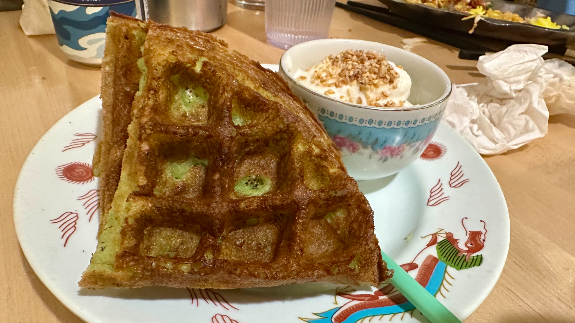 A green-tinged Pandan waffle with bowl of coconut whip and accompanying strawlike spoon on a plate.