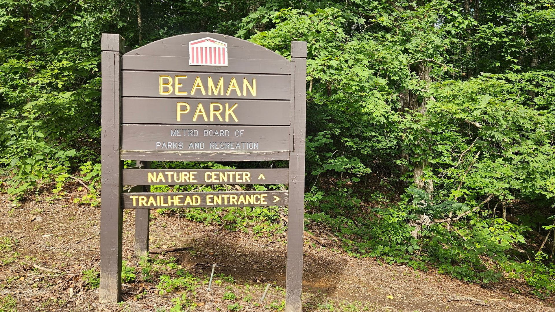 Wooden Beaman Park sign with yellow lettering sits among dense green foliage; a small emblem at the top and panels read "NATURE CENTER" and "TRAILHEAD ENTRANCE."