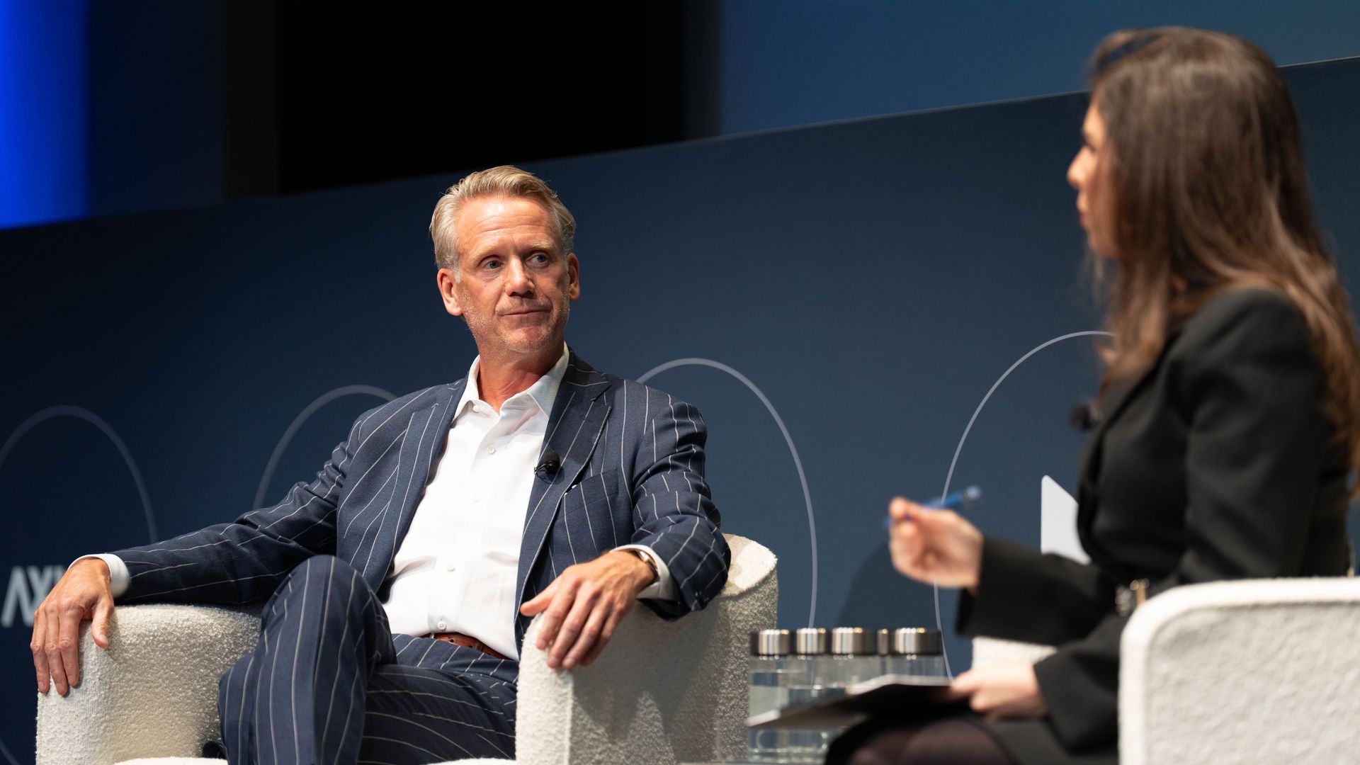 A man in a blue pinstripe suit and white shirt sits talking with a woman in a black blazer holding a pen, both seated in white chairs on a stage with a dark background.