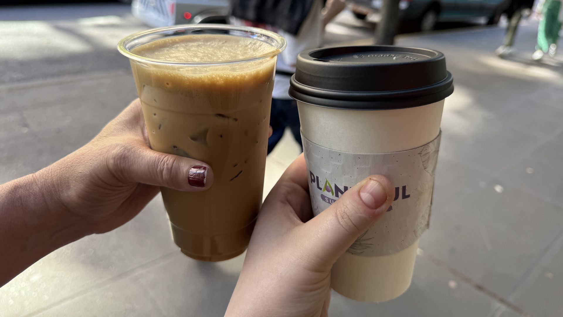 Two hands holding coffee cups outdoors: left hand holds an iced coffee in a clear plastic cup, right hand holds a hot coffee in a white cup with a black lid and a printed sleeve, busy street background.