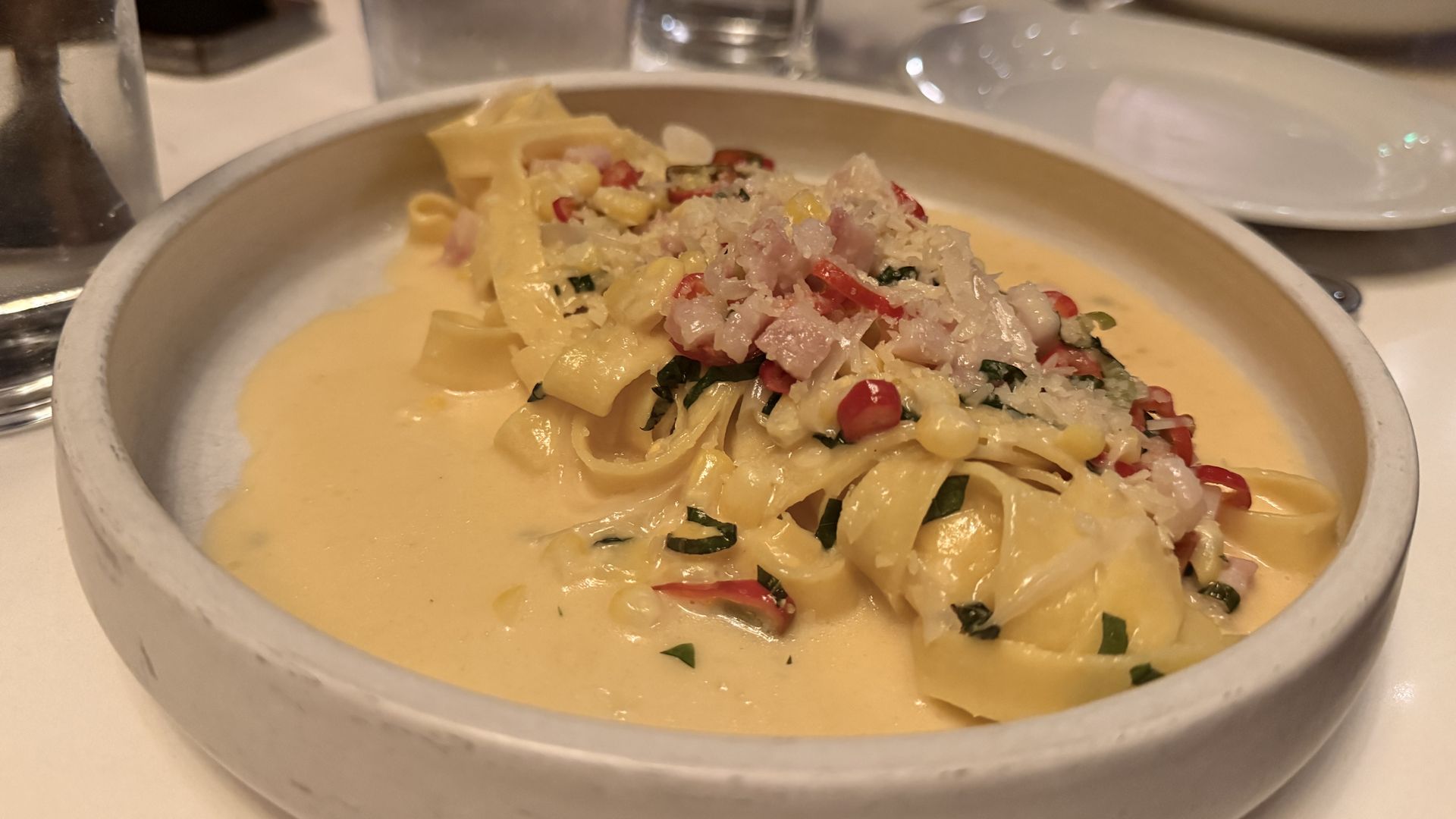 Creamy pasta in a white bowl with flat noodles, red peppers, herbs, and grated cheese; blurred glasses and plates on a dining table in the background.