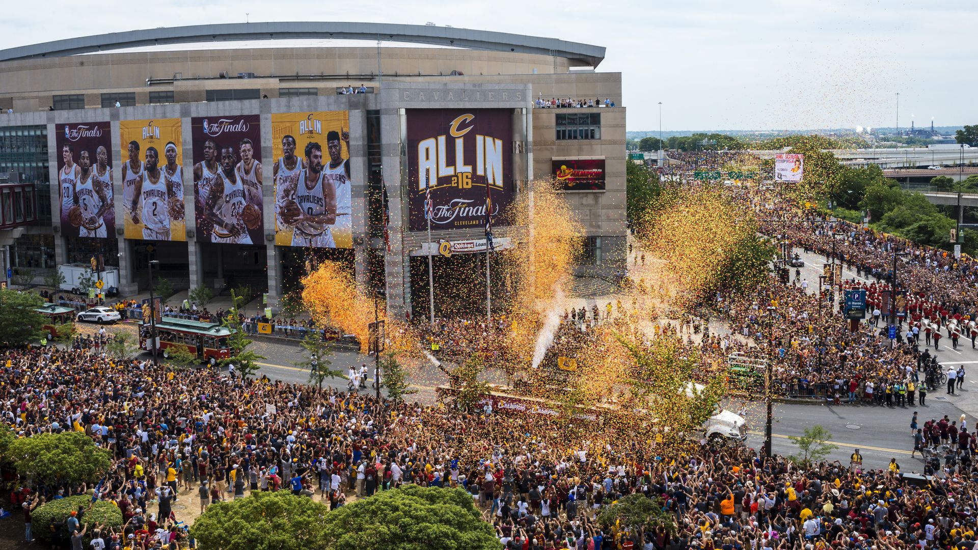 A birds eye view of the Cleveland Cavaliers victory parade in front of the basketball arena, with thousands of people lining a street and confetti being blown into the air. 