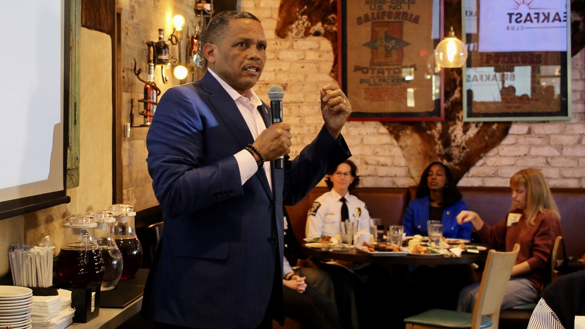 Man in navy blue suit speaking into a microphone in a rustic restaurant with exposed brick walls and people seated at tables behind him.