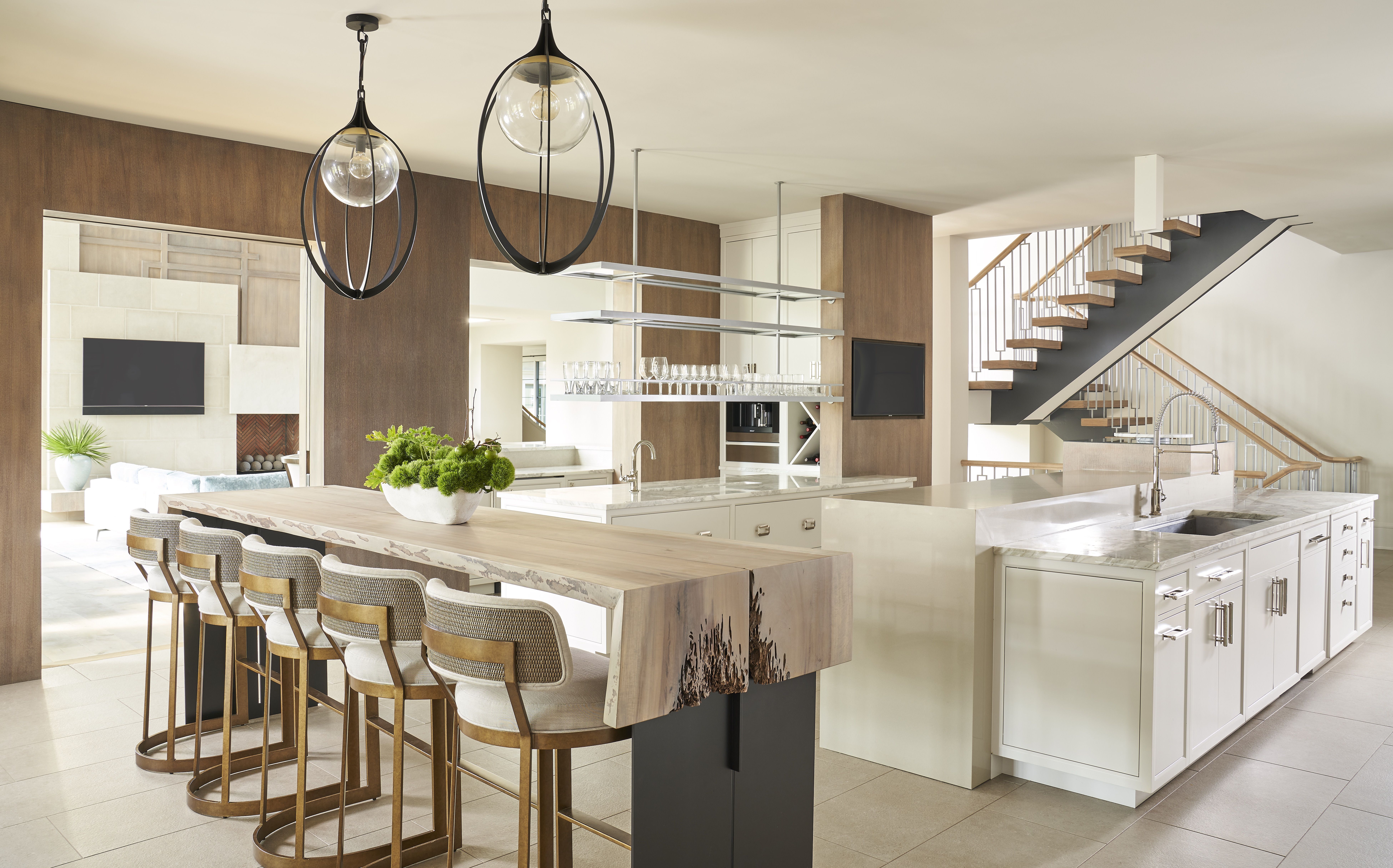 Bright modern kitchen with a long island, beige marble countertop, row of woven bar stools, hanging glass globe lights with black rings, open shelves, and a wooden staircase with metal railing.