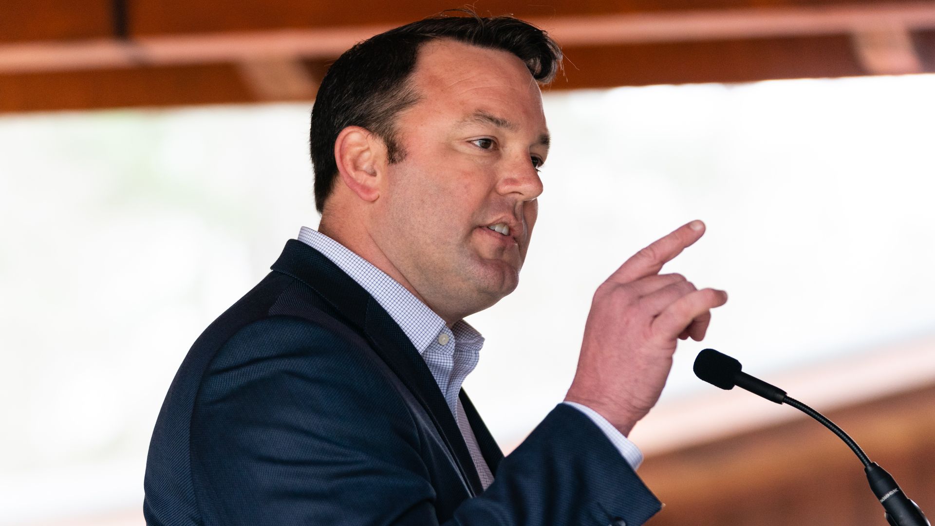 An individual wearing a suit and standing at a microphone speaks to a crowd located outside the frame