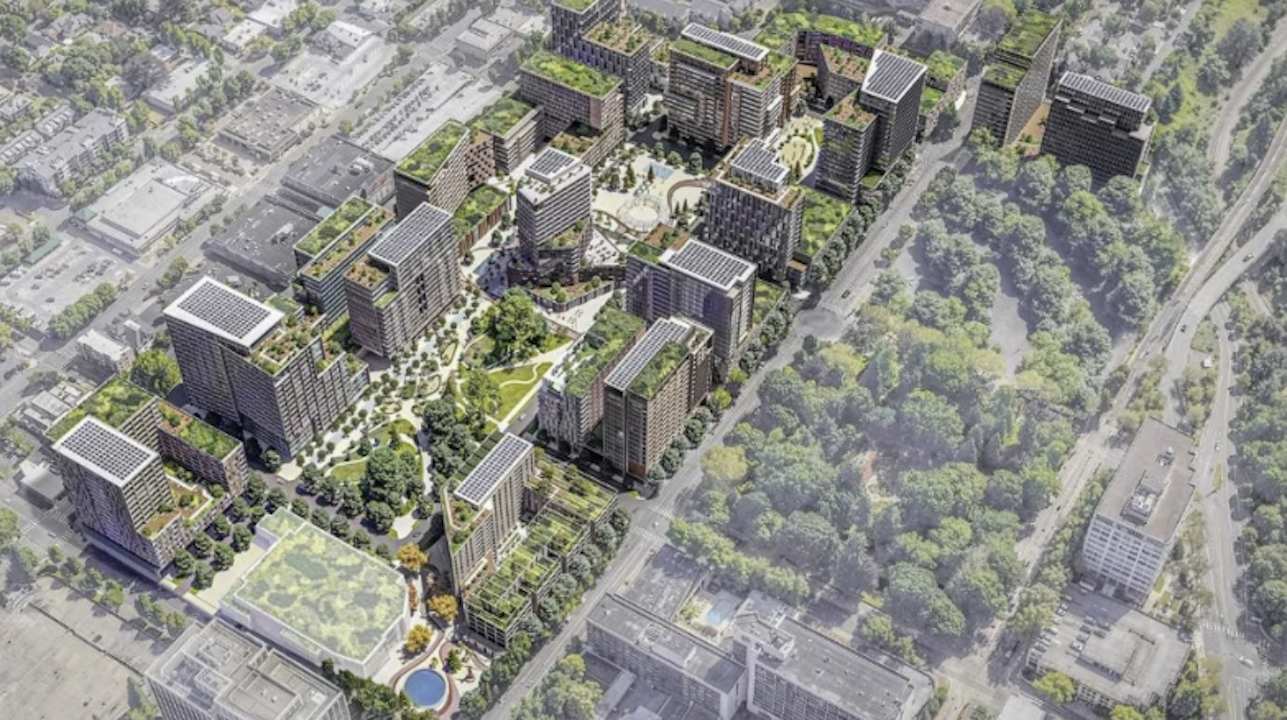 Aerial view of a modern residential development: clustered high-rises surround a central park with winding paths, green roofs, and courtyards; tree-lined streets and a nearby forest.