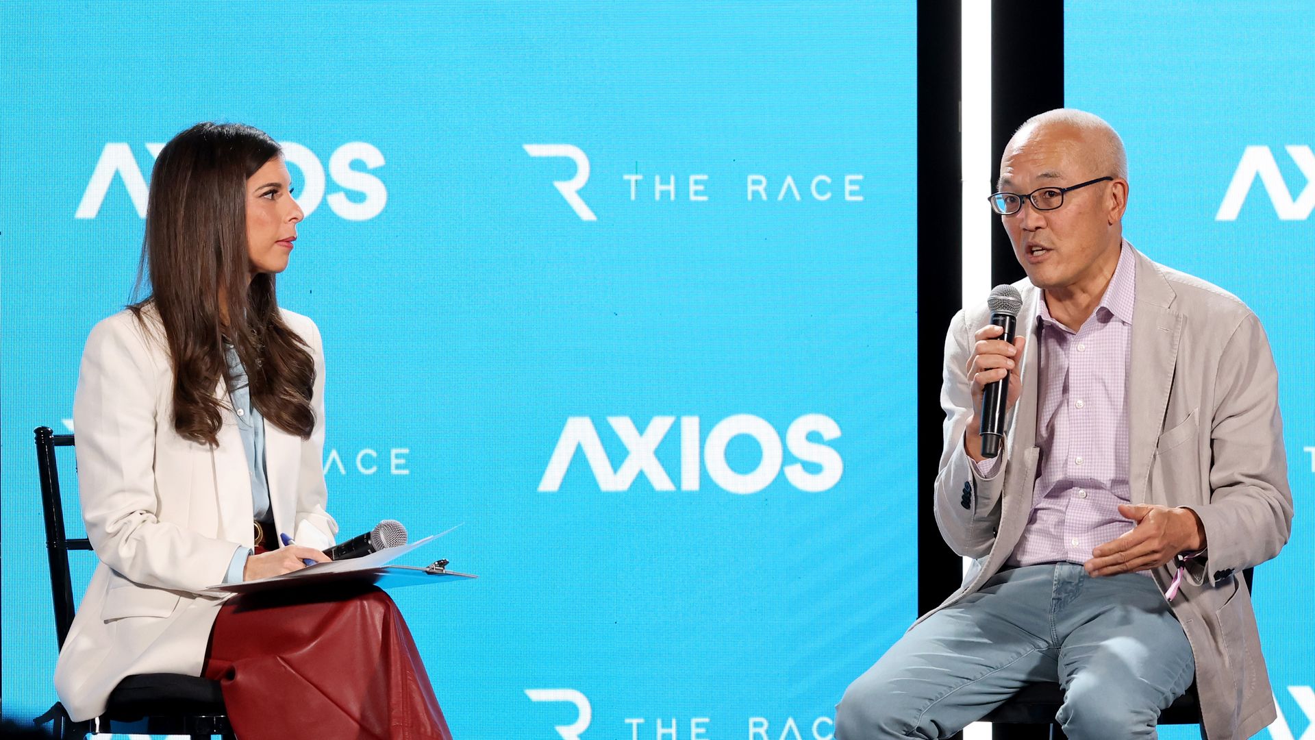 Two panelists on a stage with a blue Axios backdrop; a woman in a white blazer holds a clipboard, while a man in a beige jacket speaks into a microphone.