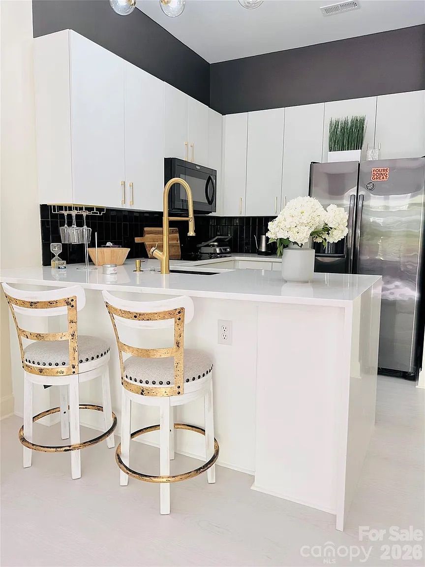 Modern kitchen with white cabinets, black backsplash, gold faucet, two vintage-style bar stools with white cushions, and a stainless steel refrigerator with positive message magnet.