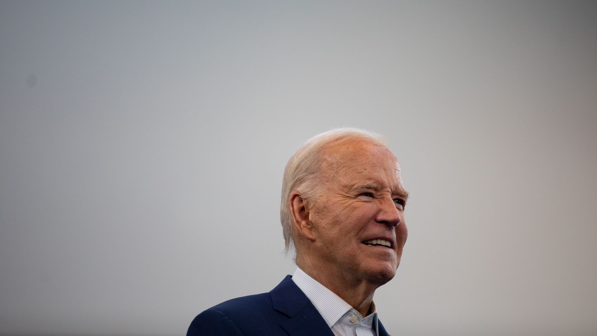 President Biden, wearing a blue suit and standing in front of a white backdrop.
