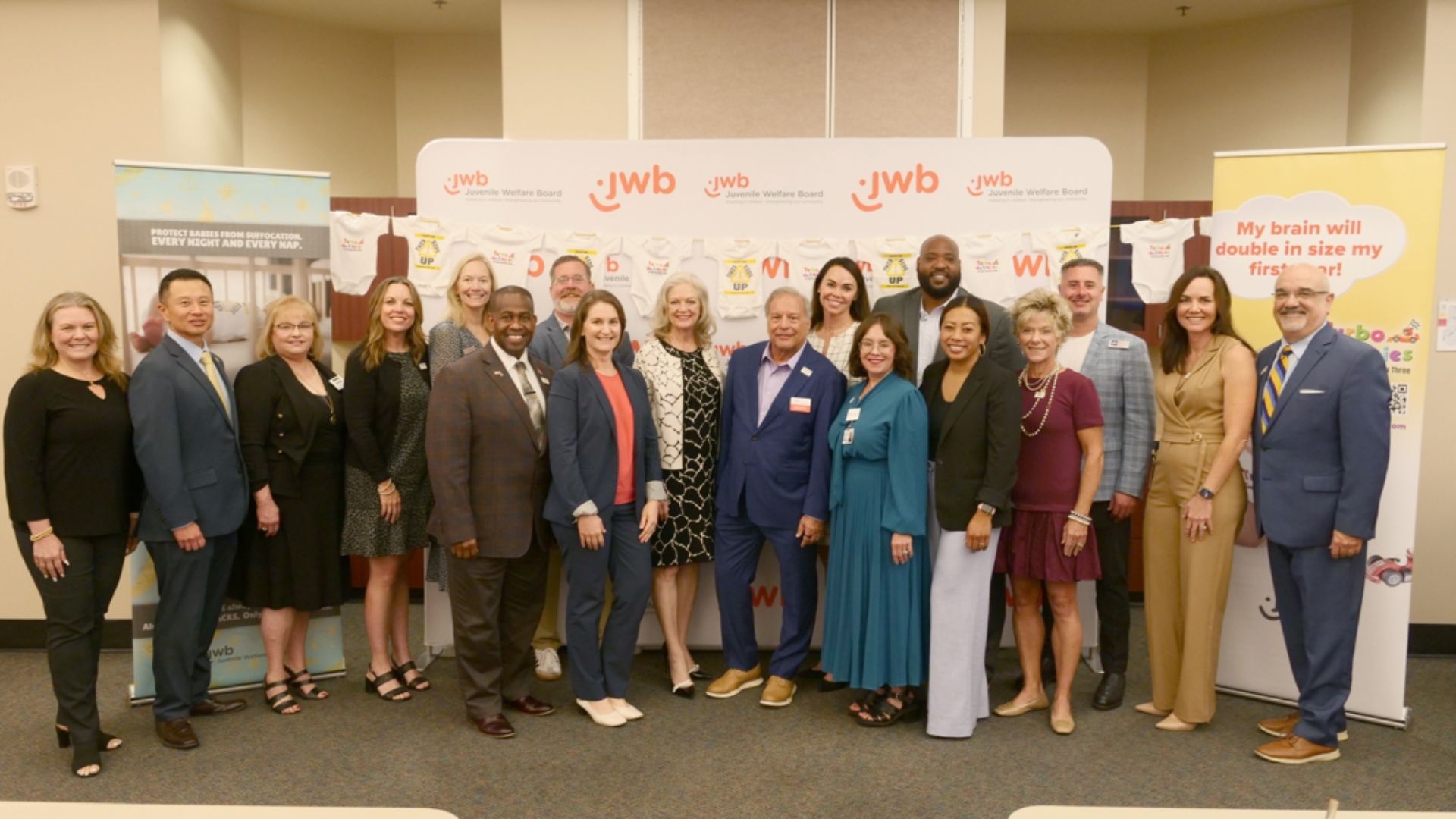 Group of sixteen diverse professionals standing indoors in front of banners and a backdrop with "JWB Juvenile Welfare Board" logos, smiling at the camera, dressed in business attire.