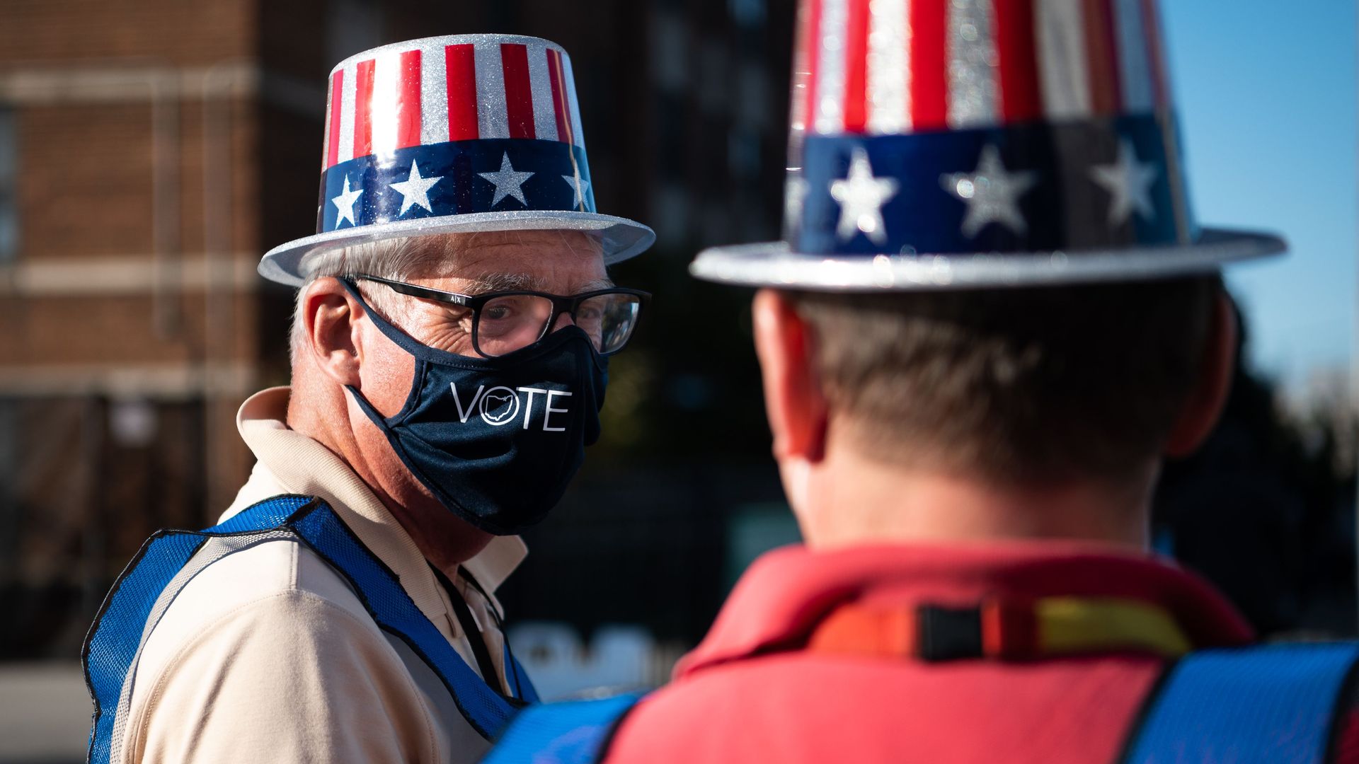 A man in an American flag-themed hat and "Vote" mask stands outside a polling place. 