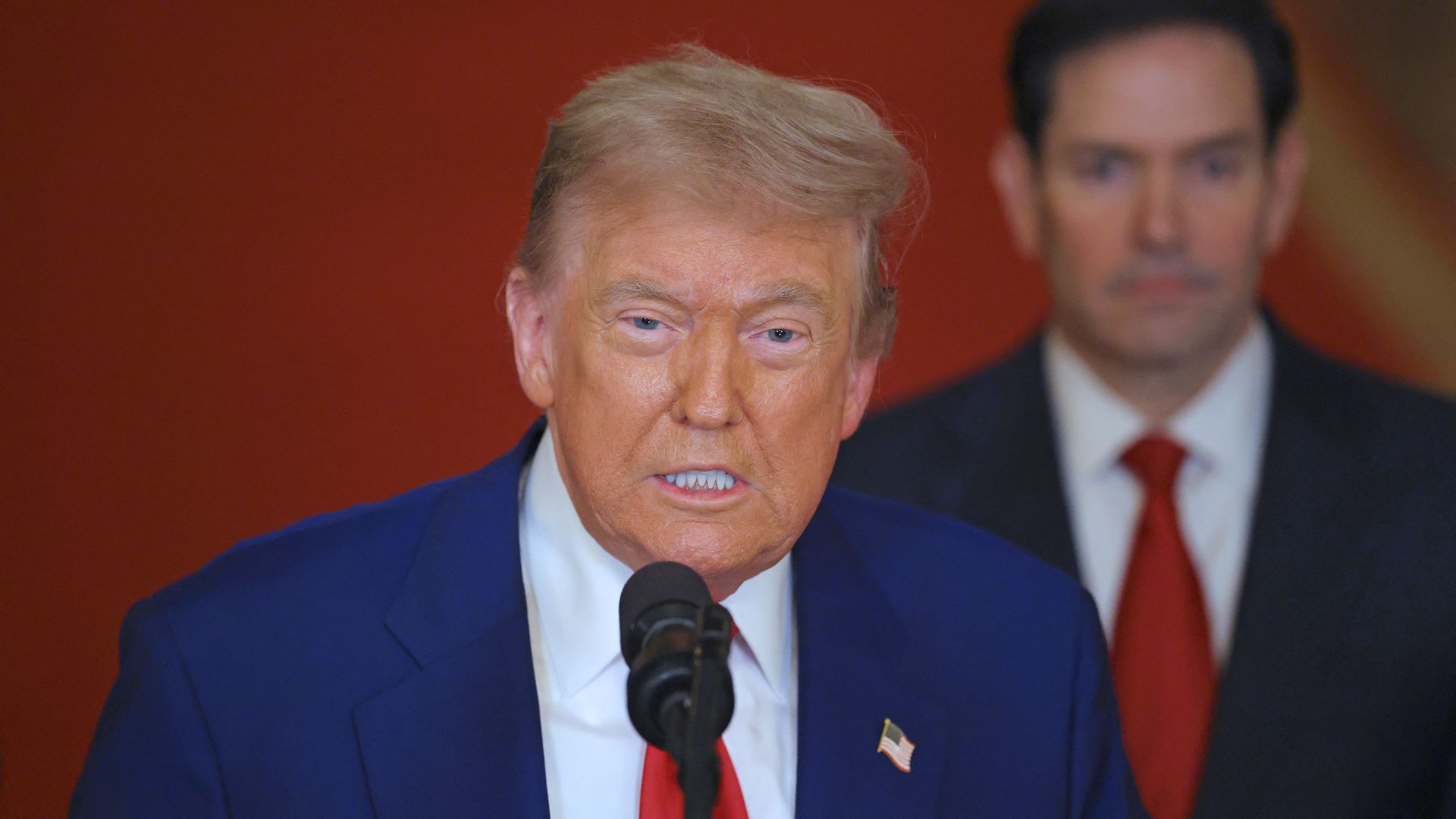 President Trump speaks into a microphone as Marco Rubio stands behind him.