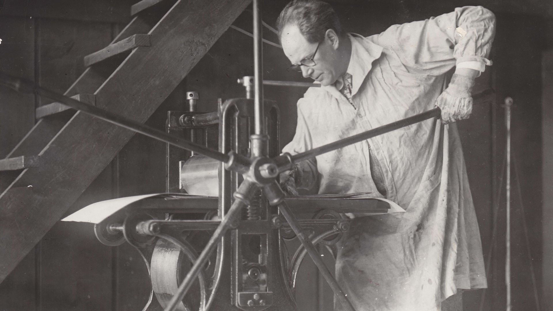 Black-and-white photo of a man in a light lab coat and gloves, wearing glasses, operating a vintage metalworking machine. He leans over a bench with gears and a vertical rod, wooden beams in the background.