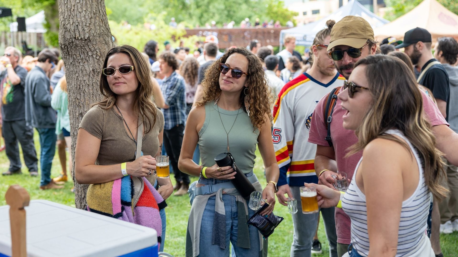 Group of friends at an outdoor festival. Three women wearing sunglasses stand by a blue cooler, holding drinks, as a crowd chats in the grassy park.