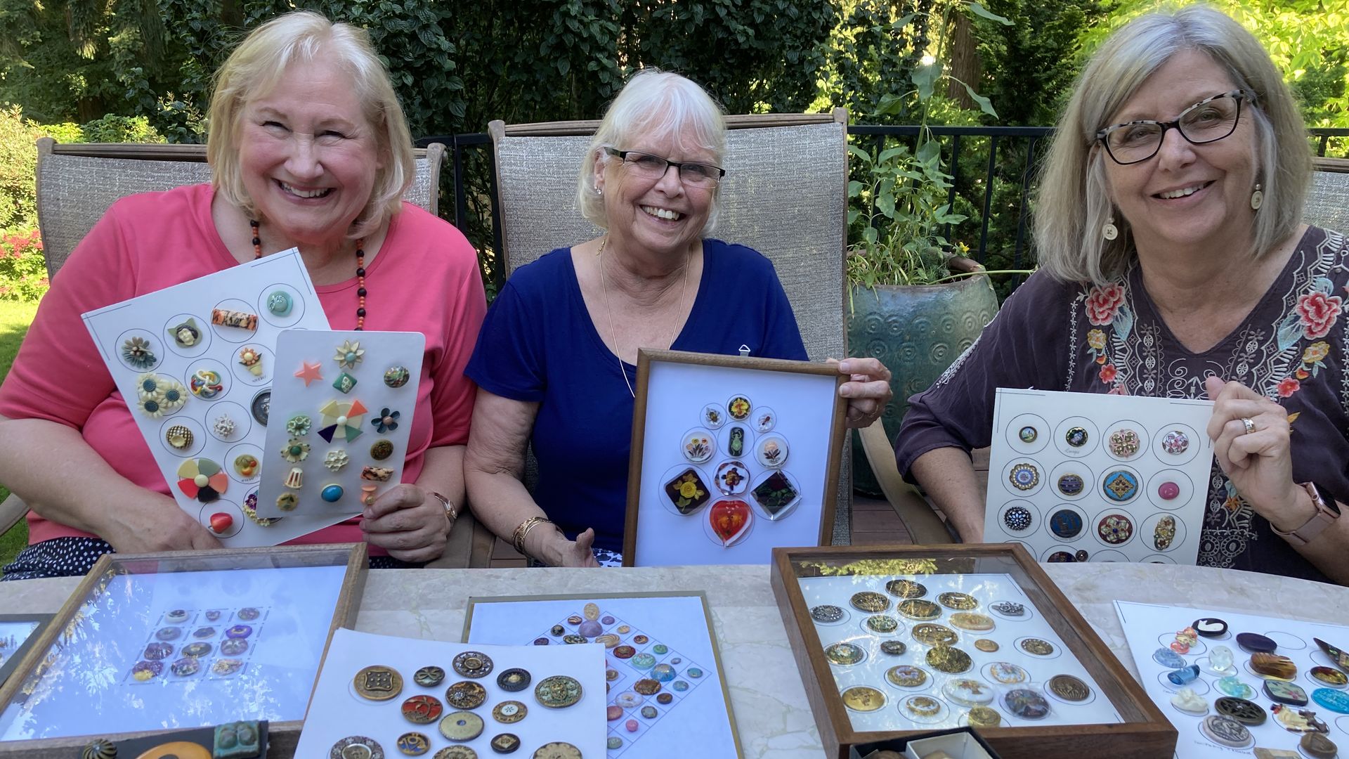 Three women smile as they hold up displays of collectible buttons on white cardstock.