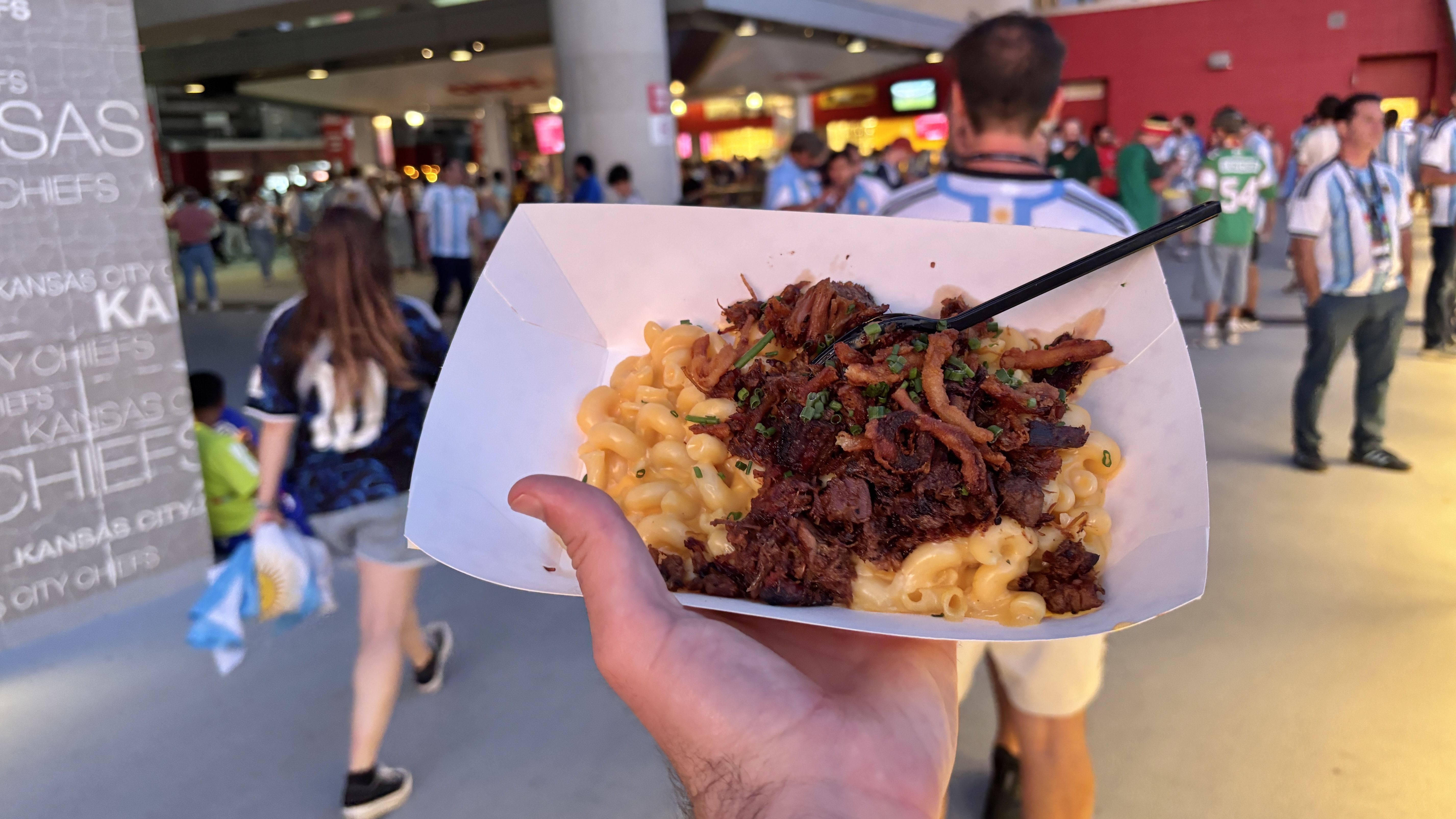 Close-up of a hand holding a white tray of macaroni and cheese topped with crispy bacon bits and chives, with a crowd of fans in blue-and-white jerseys in the background.