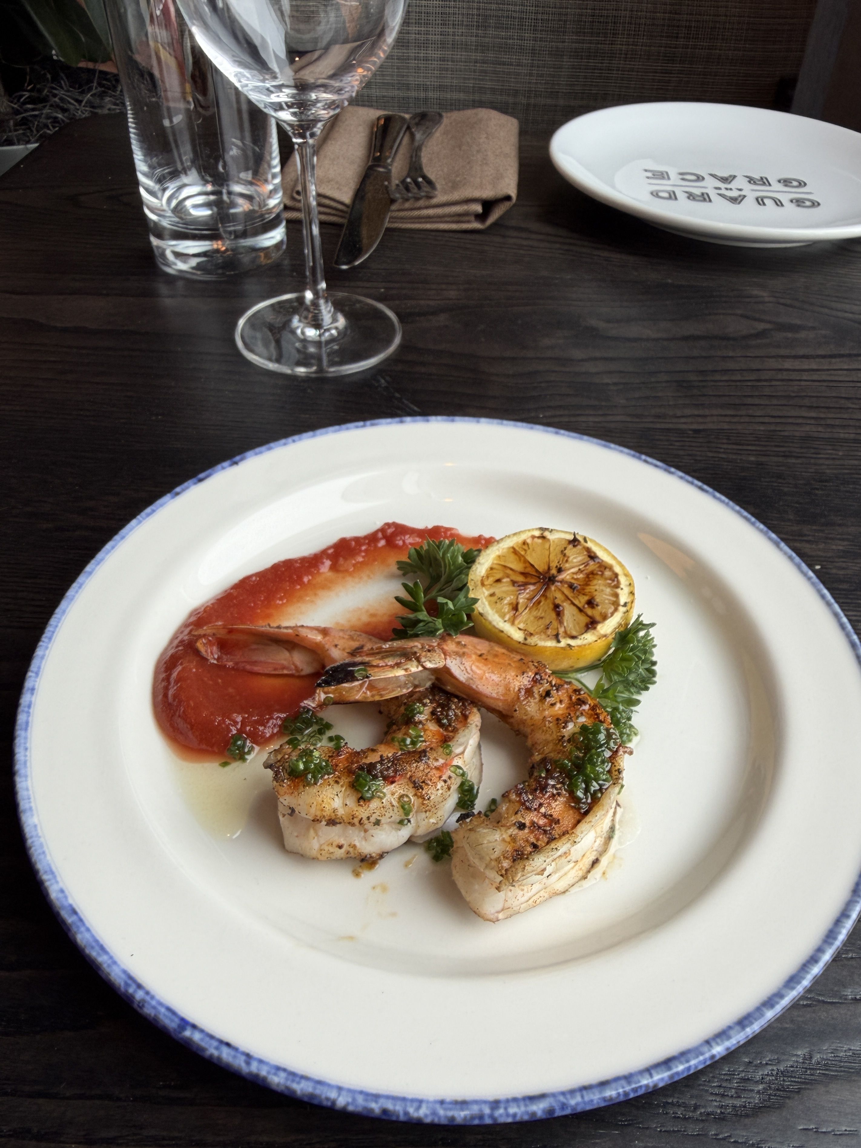 Plate of grilled shrimp with lemon half, parsley, and red sauce on a dark wooden table. Nearby: a wine glass, a water glass, and a folded napkin with cutlery.