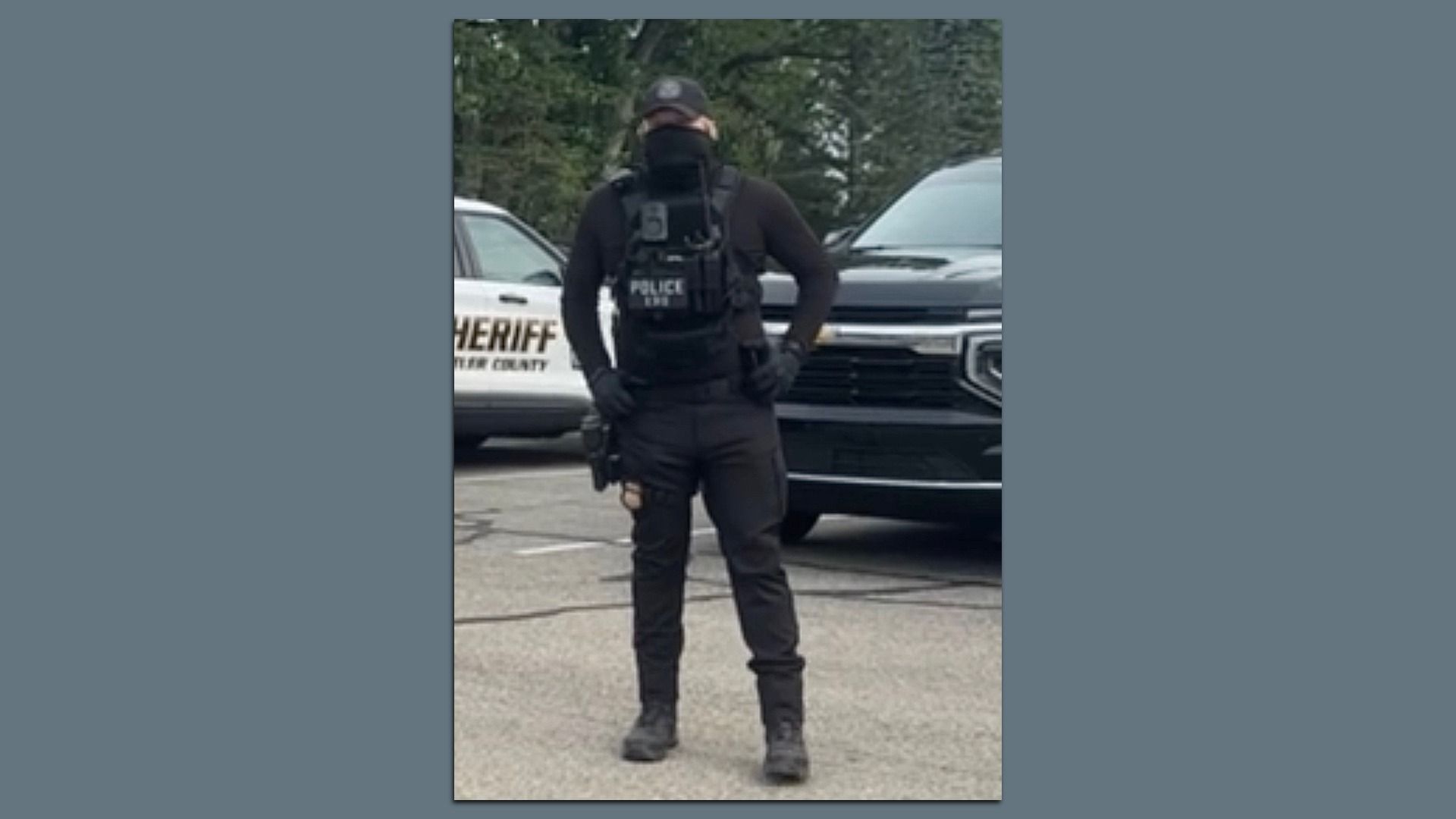 Masked, black-clad police officer in tactical gear and a bulletproof vest stands in a parking lot; behind him are a sheriff's patrol car and a gray pickup truck.