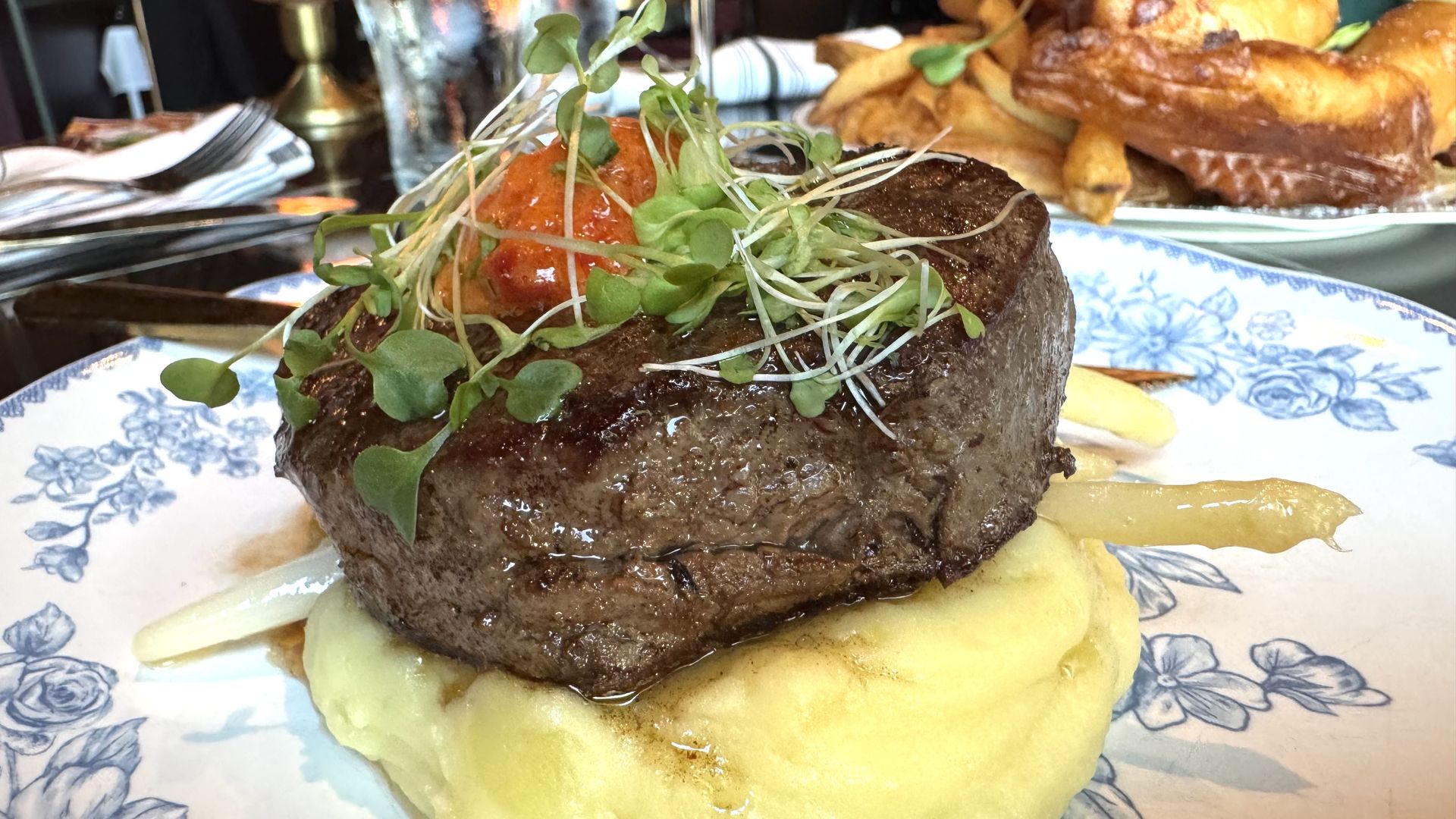 Thick, seared steak topped with microgreens and a small relish, plated on creamy mashed potatoes on a blue floral dish; background shows more food.