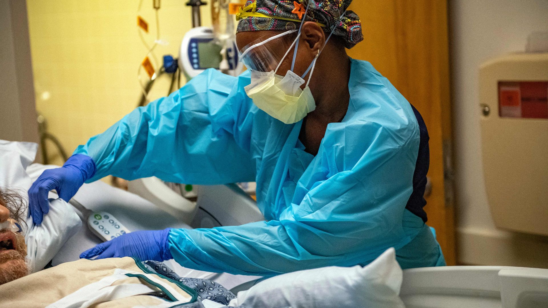Photo of a masked nurse in scrubs adjusting a pillow for an patient in a bed in the ICU ward