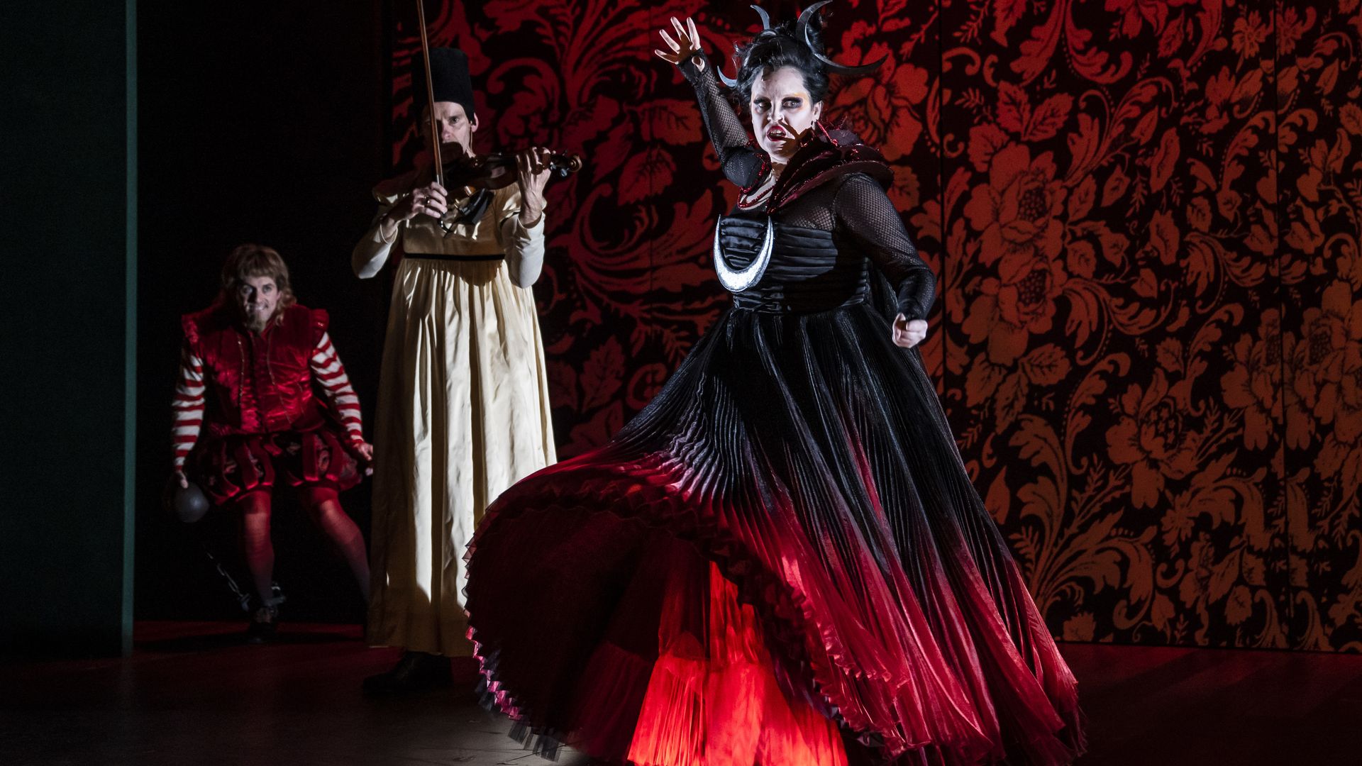 Actor in black and red long dress with her arm high in the air and two characters behind her in a stage production.