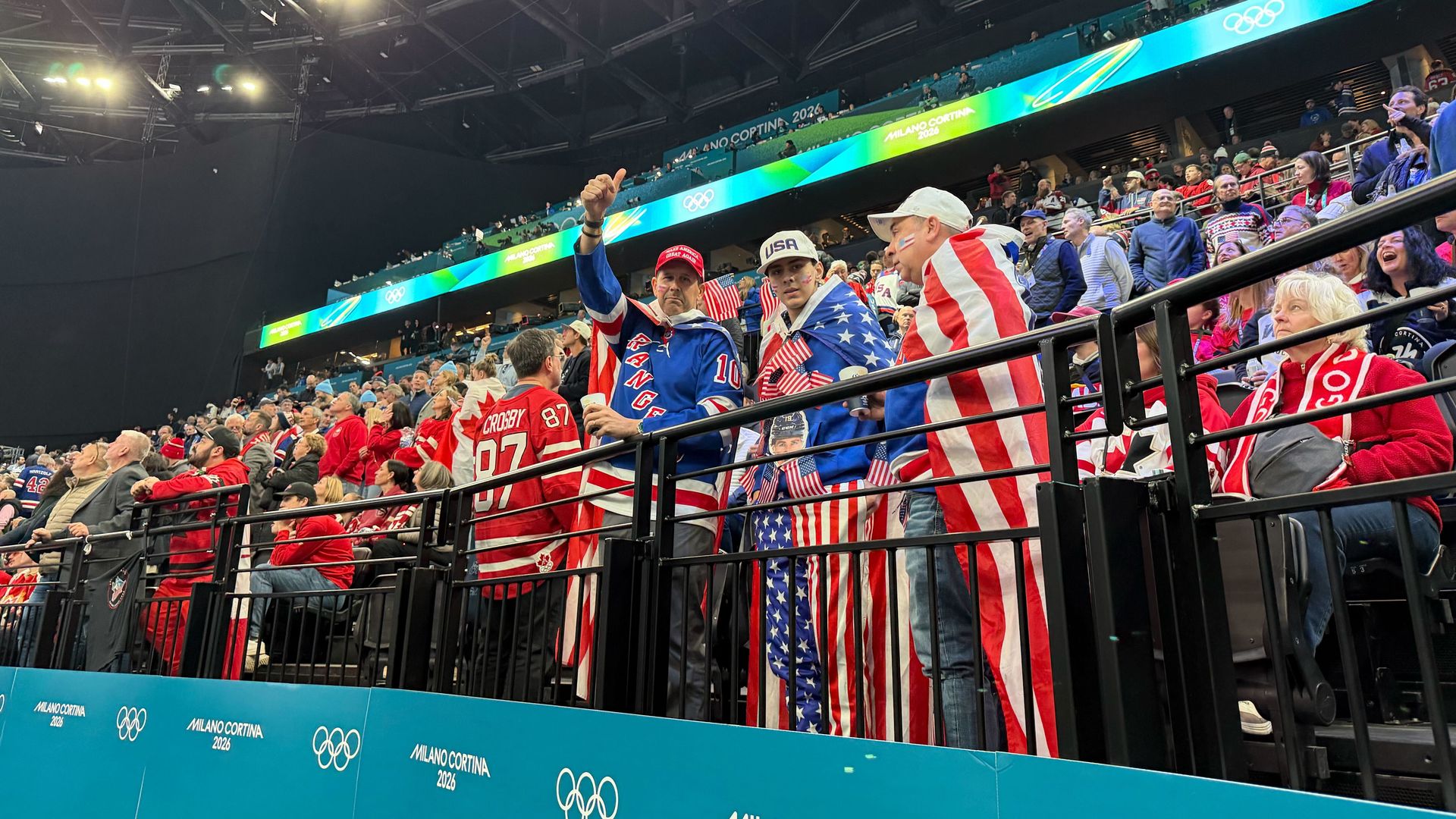 Crowd in an arena at Milano Cortina 2026 Olympics, fans dressed in USA and Canada colors, flags, and jerseys, with bright arena lights overhead and Olympic logos visible.