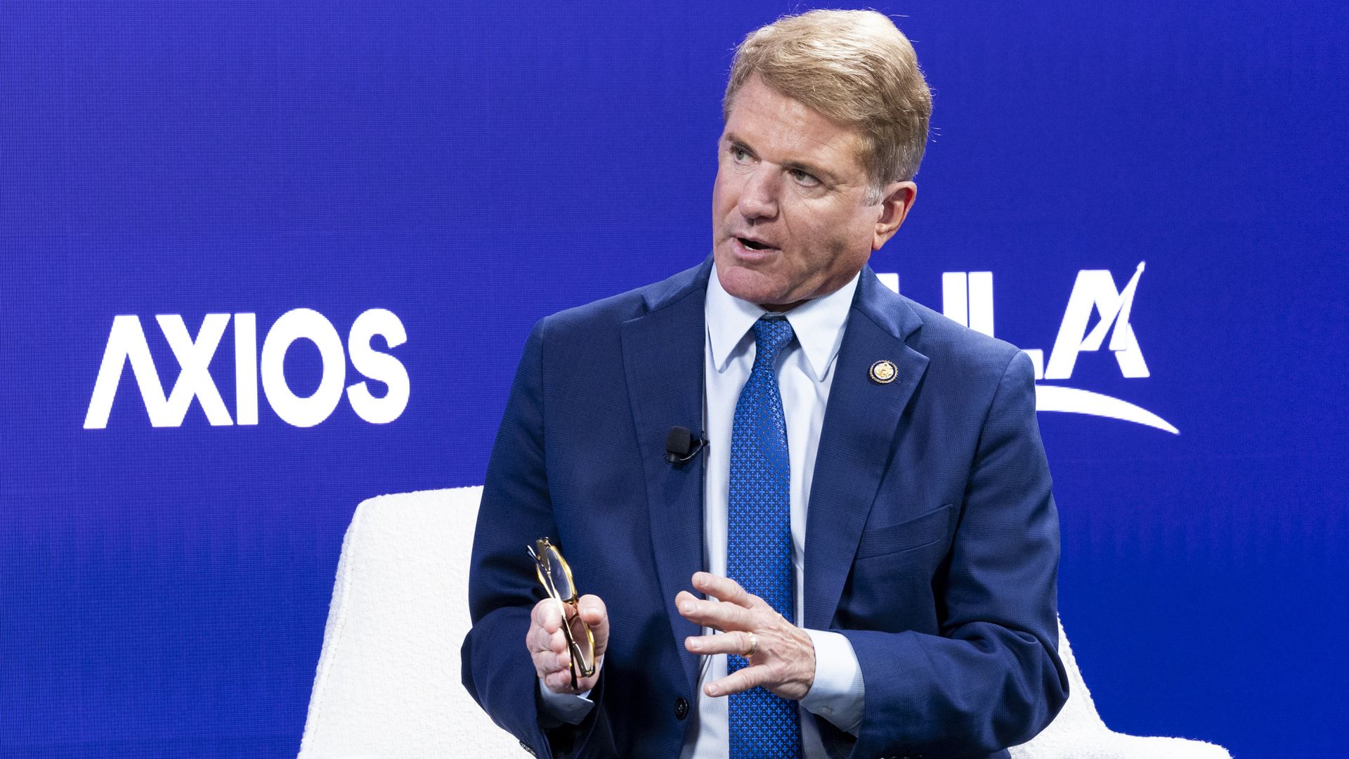 Man in blue suit and tie seated on white chair speaking, holding glasses, with blue background featuring white "AXIOS" text.