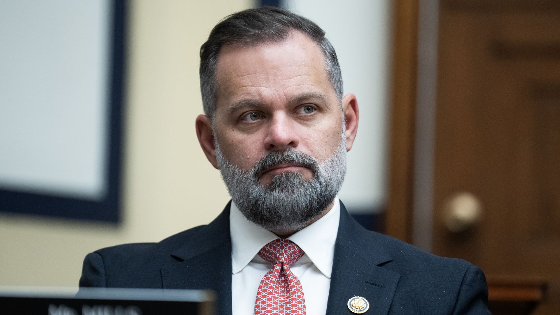 A bearded man in a dark suit and red tie sits in a courtroom, looking to the left with a thoughtful expression. A lapel badge is visible; blurred wood panel background.