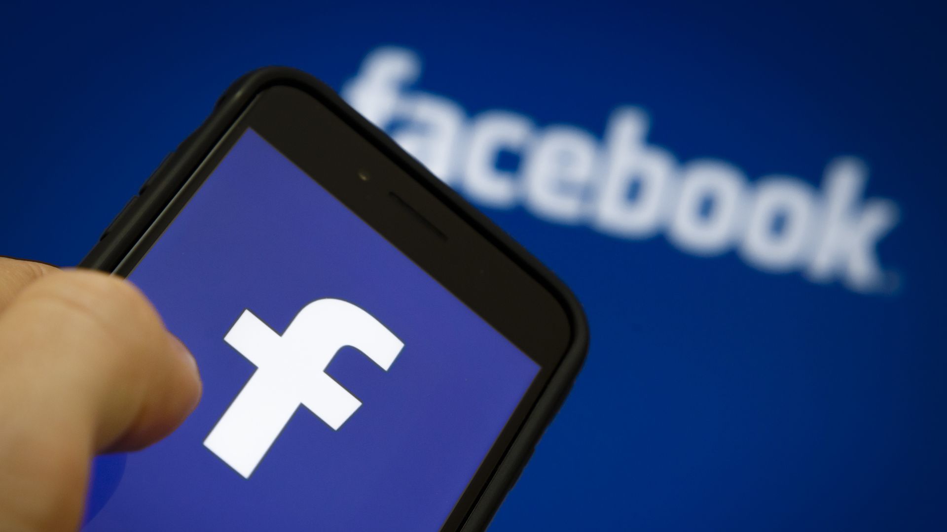a thumb pressed on a smart phone displaying the Facebook logo