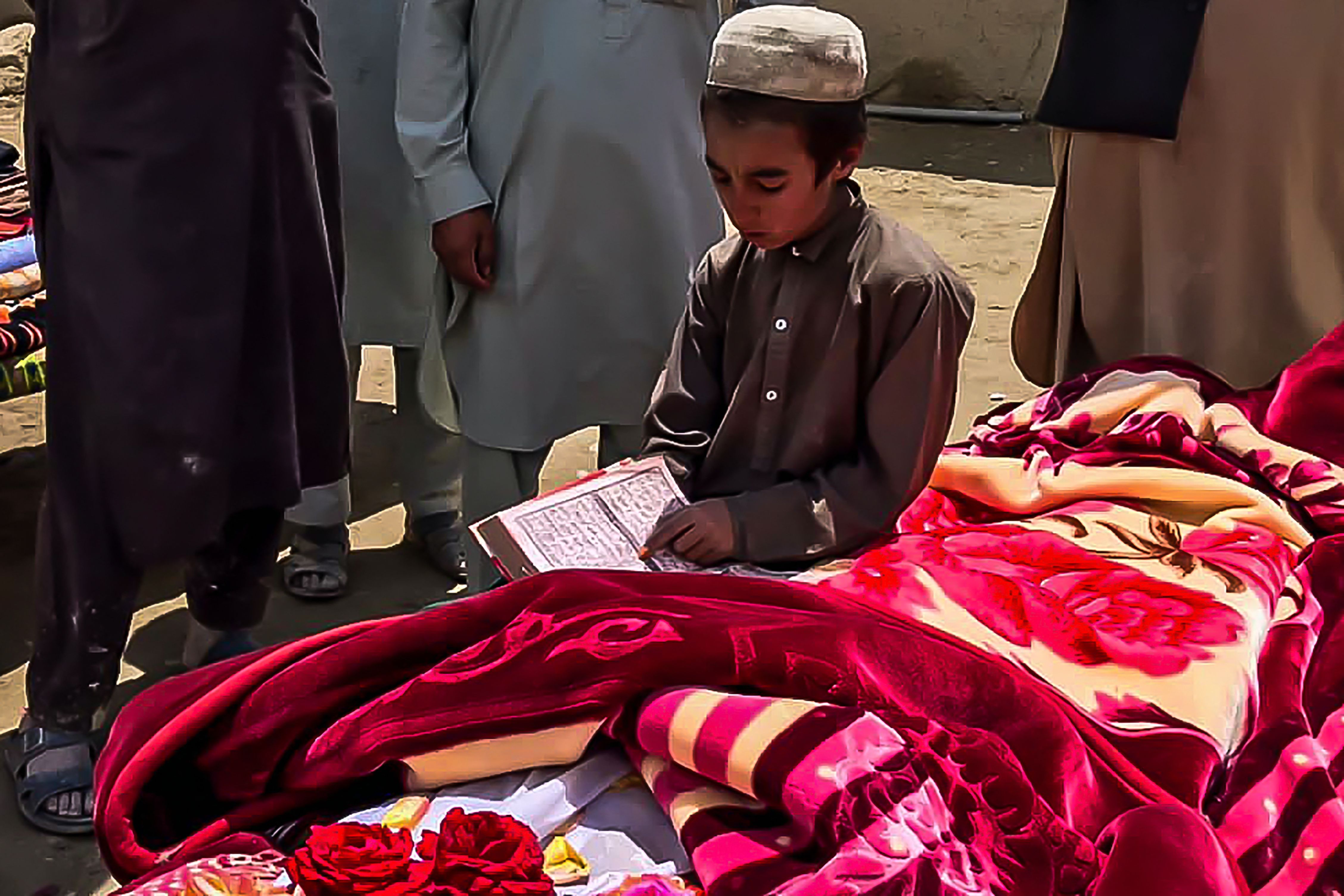 A boy reads from a religious text over a dead body covered in a blanket. 