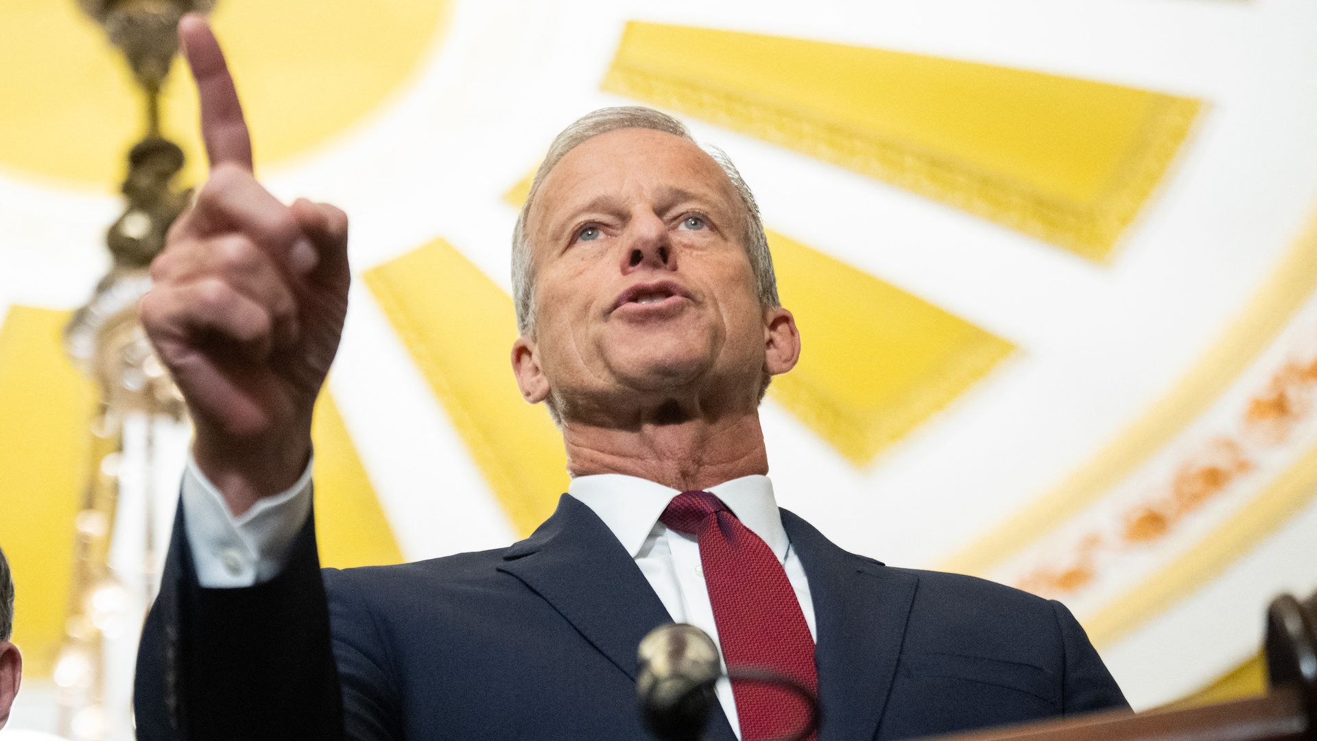 John Thune in a dark suit and red tie speaking passionately behind a podium with a microphone, raising one finger, with a yellow and white decorative ceiling background.