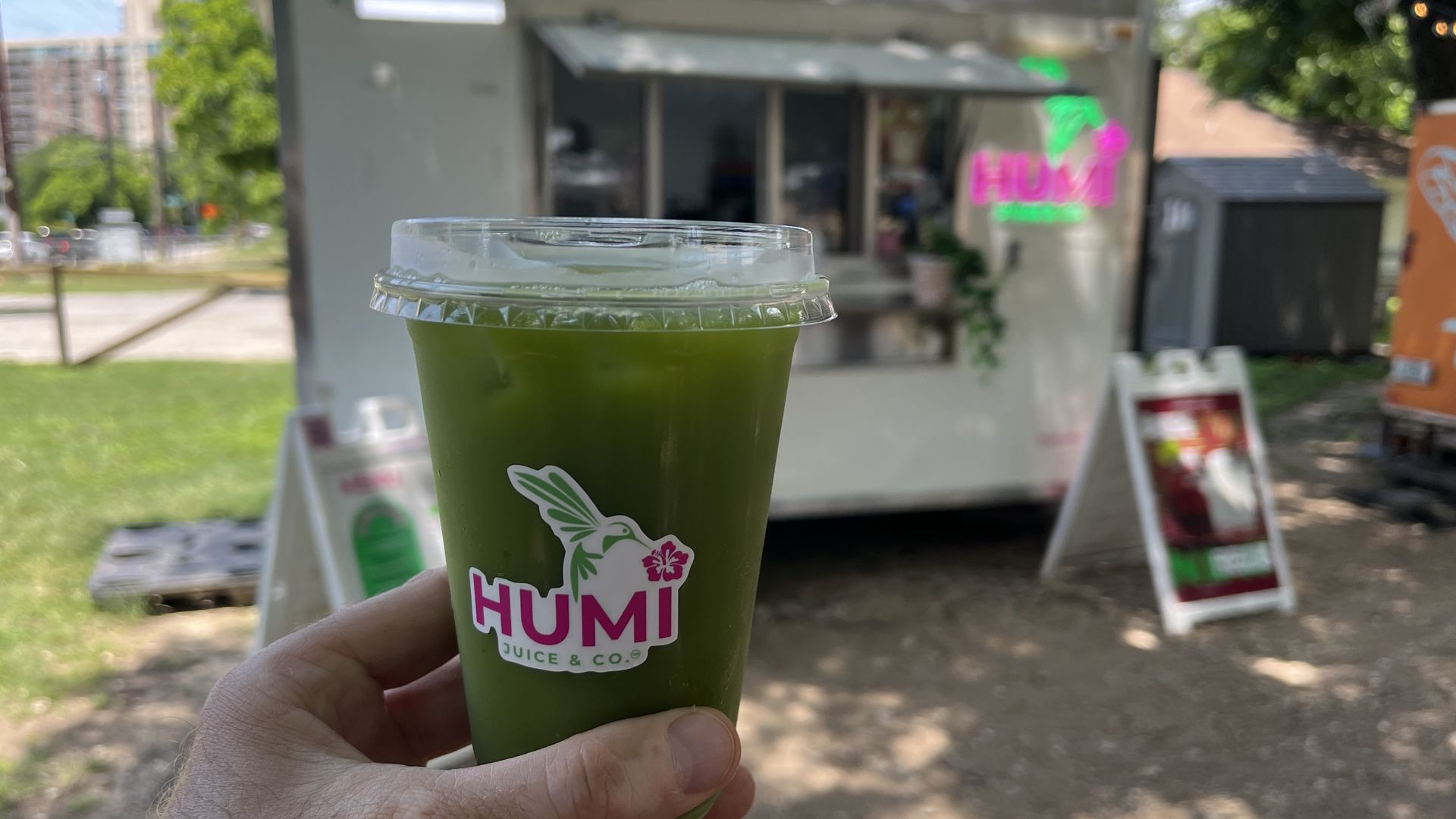 Close-up of a green juice cup with a clear lid held by a hand; HUMI logo with a bird and hibiscus visible. Blurred background shows a gray food truck and neon pink-green sign in a sunny park.