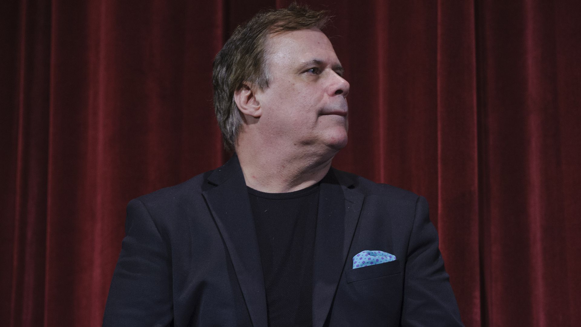 A middle-aged man in a dark blazer and black shirt sits on a stage with a red velvet curtain behind; a blue pocket square in his jacket and a microphone in front, looking to the right.