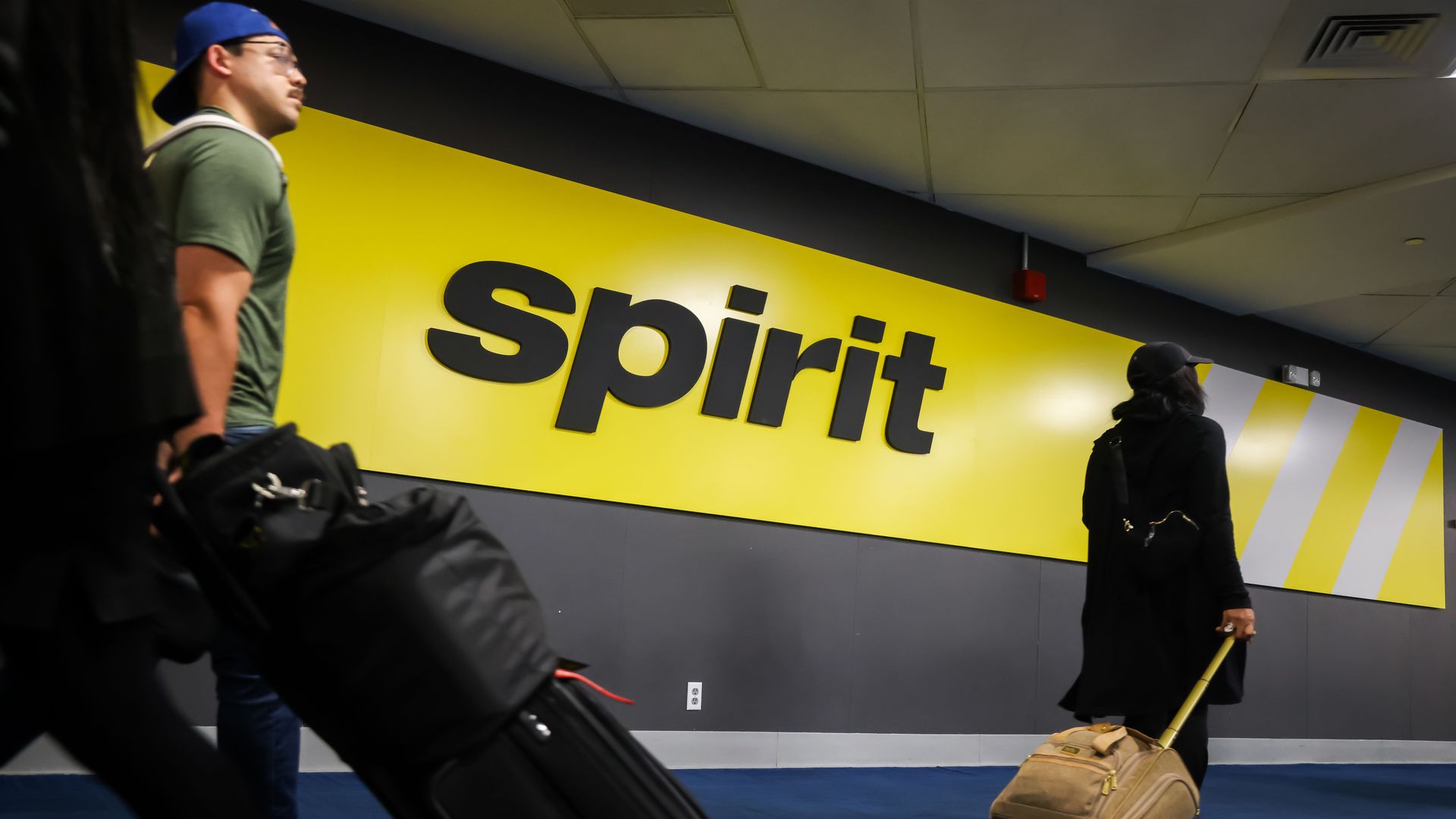 Two travelers pulling suitcases walk past a bright yellow wall with large black letters spelling "spirit" inside an airport terminal.