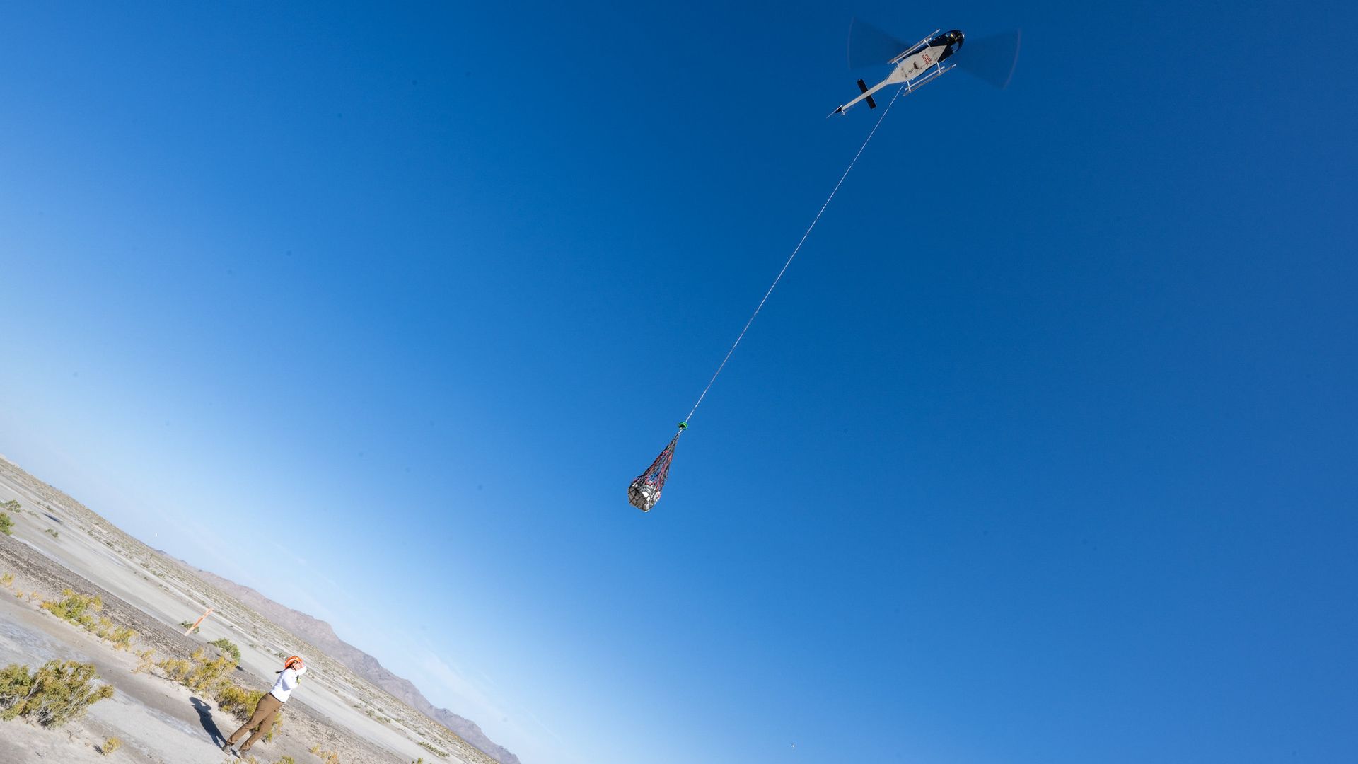 A person looks on as a helicopter raises a capsule carrying an asteroid sample into a clear blue sky above Utah