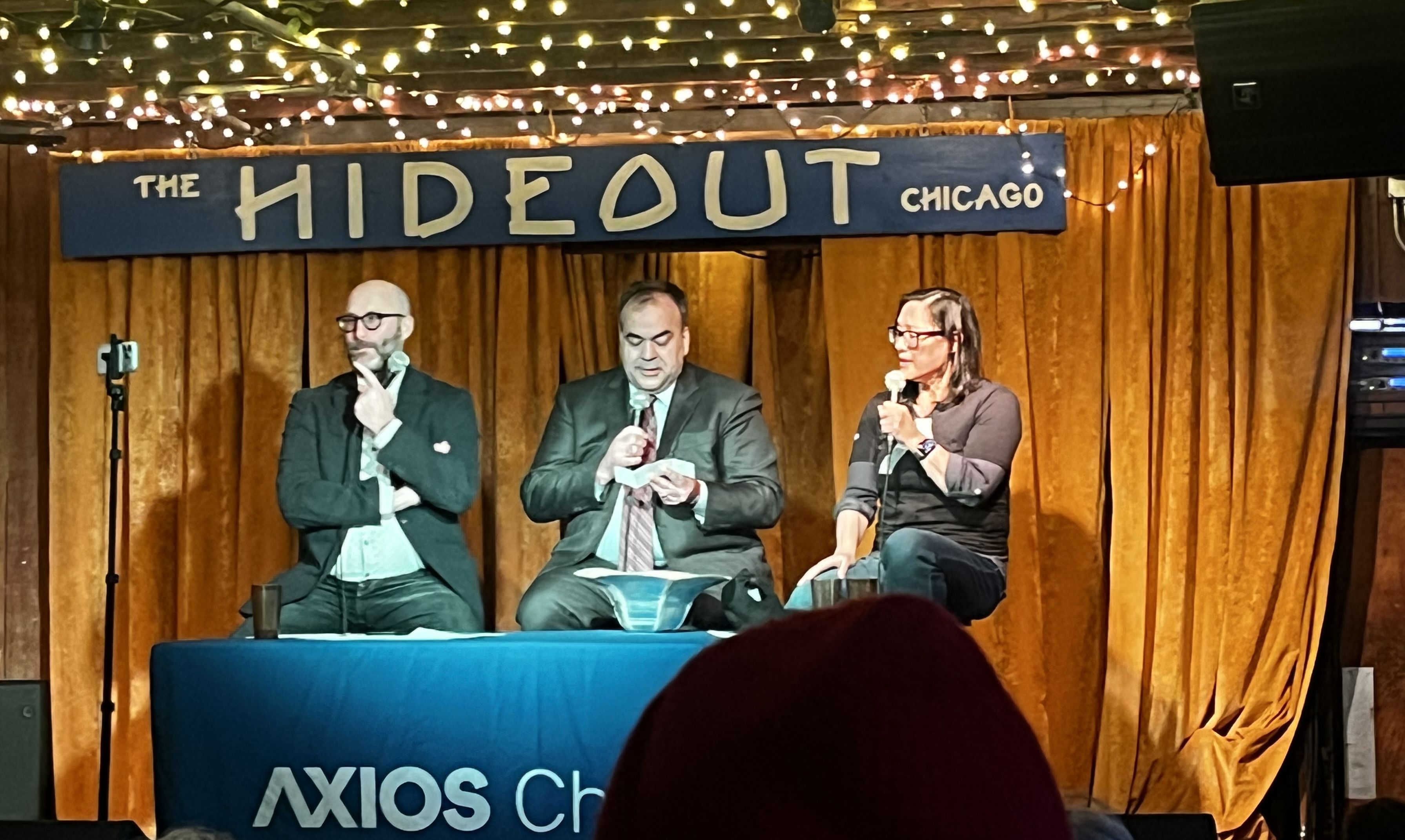 Panel at The Hideout Chicago. Three speakers behind a blue table under string lights and gold curtains; left: bearded man with glasses, middle: man reading notes, right: woman with microphone.