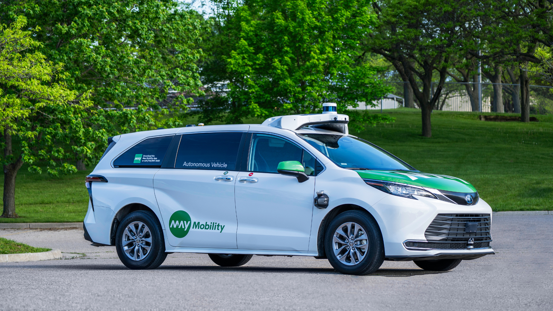 White and green autonomous vehicle with "MAY Mobility" logo parked on road, equipped with sensors and cameras, surrounded by green trees and grass in the background.