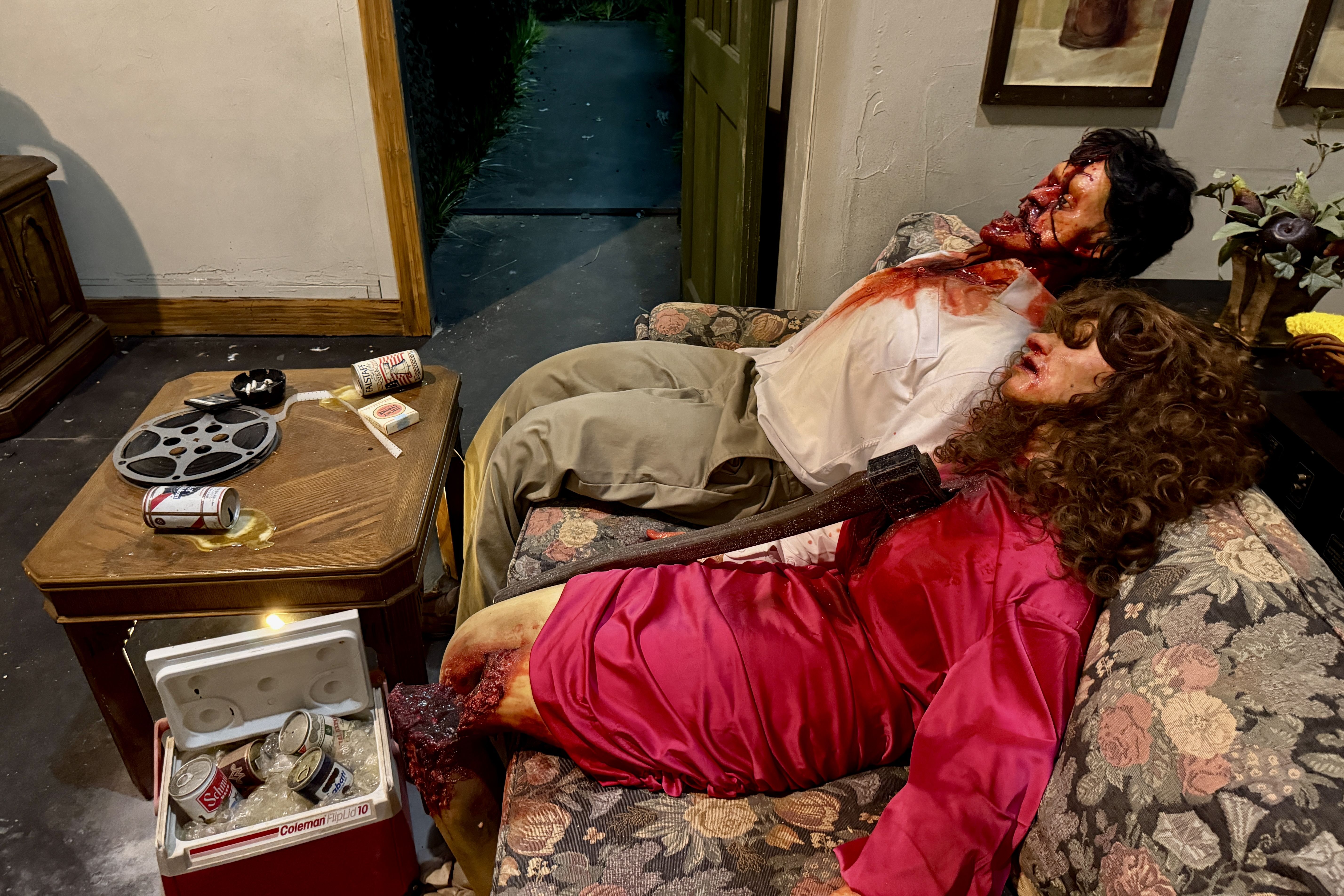 Two bloodied people slump on a floral sofa; one has long black hair, the other a white shirt stained red. A coffee table holds cans and a film reel; an open door reveals a dark outdoor path.