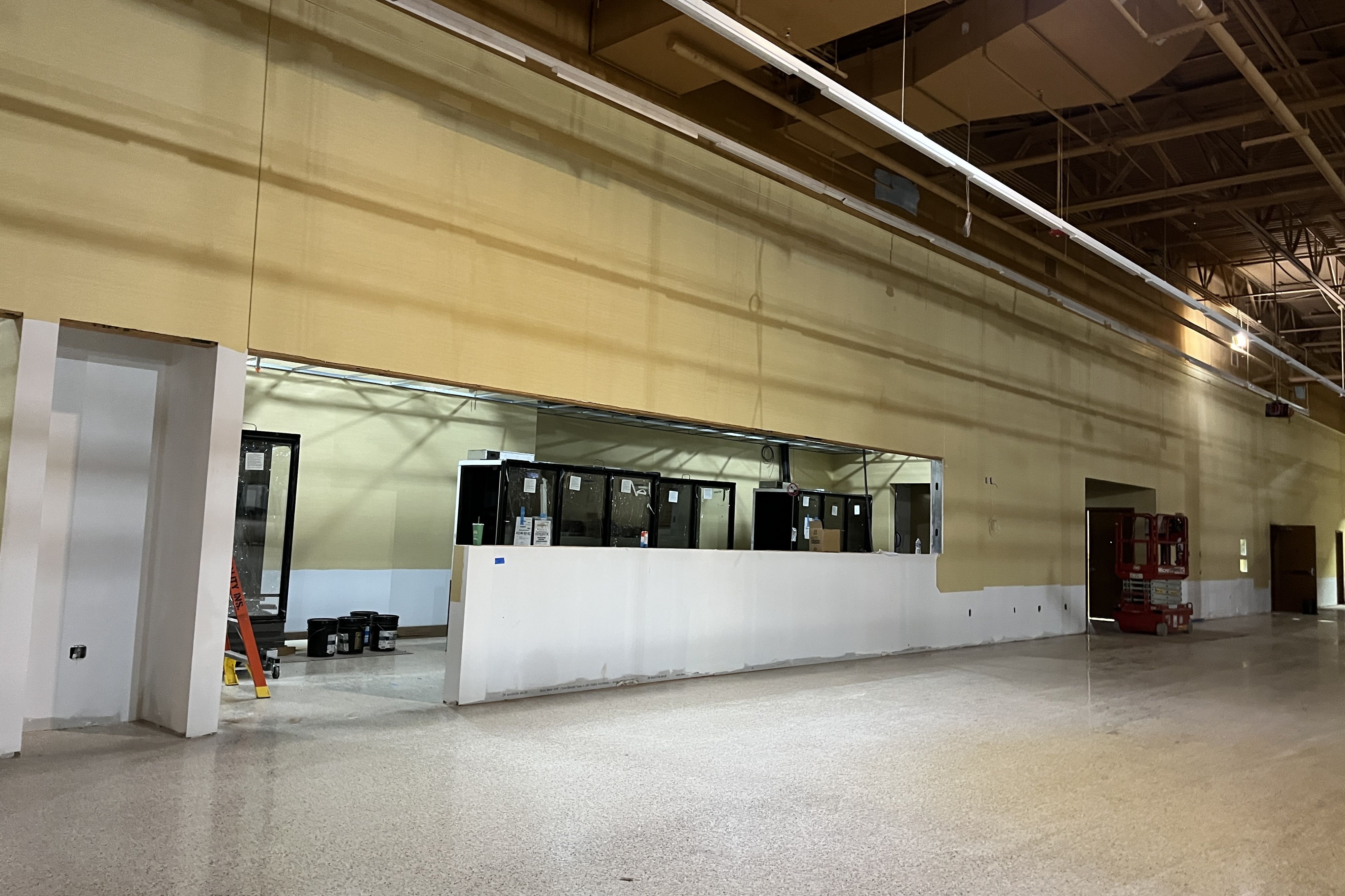 Interior of a large unfinished showroom: beige walls, white front desk, glass display cases, exposed ceiling pipes, and a yellow ladder with a red lift in the background.
