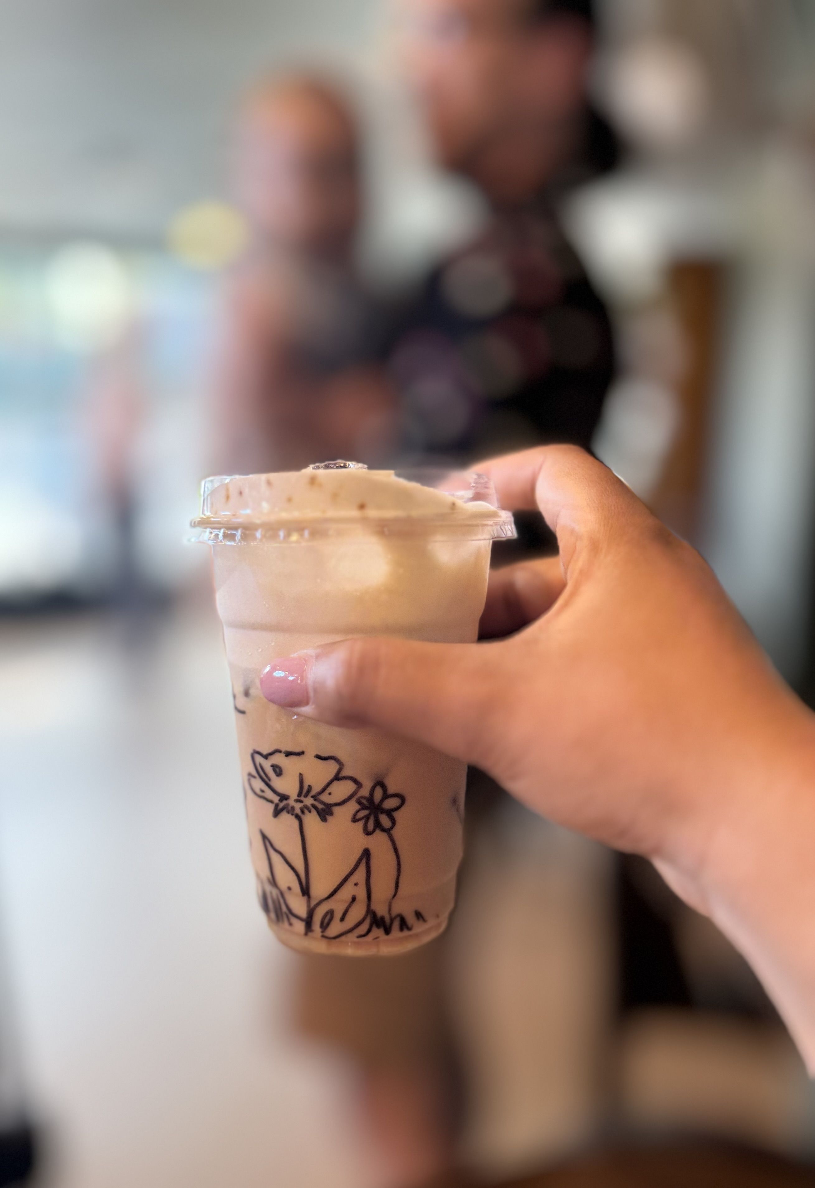 Close-up of a hand with pink nails gripping a clear plastic cup with a clear lid, iced beverage with floral drawings; blurred people in the background.