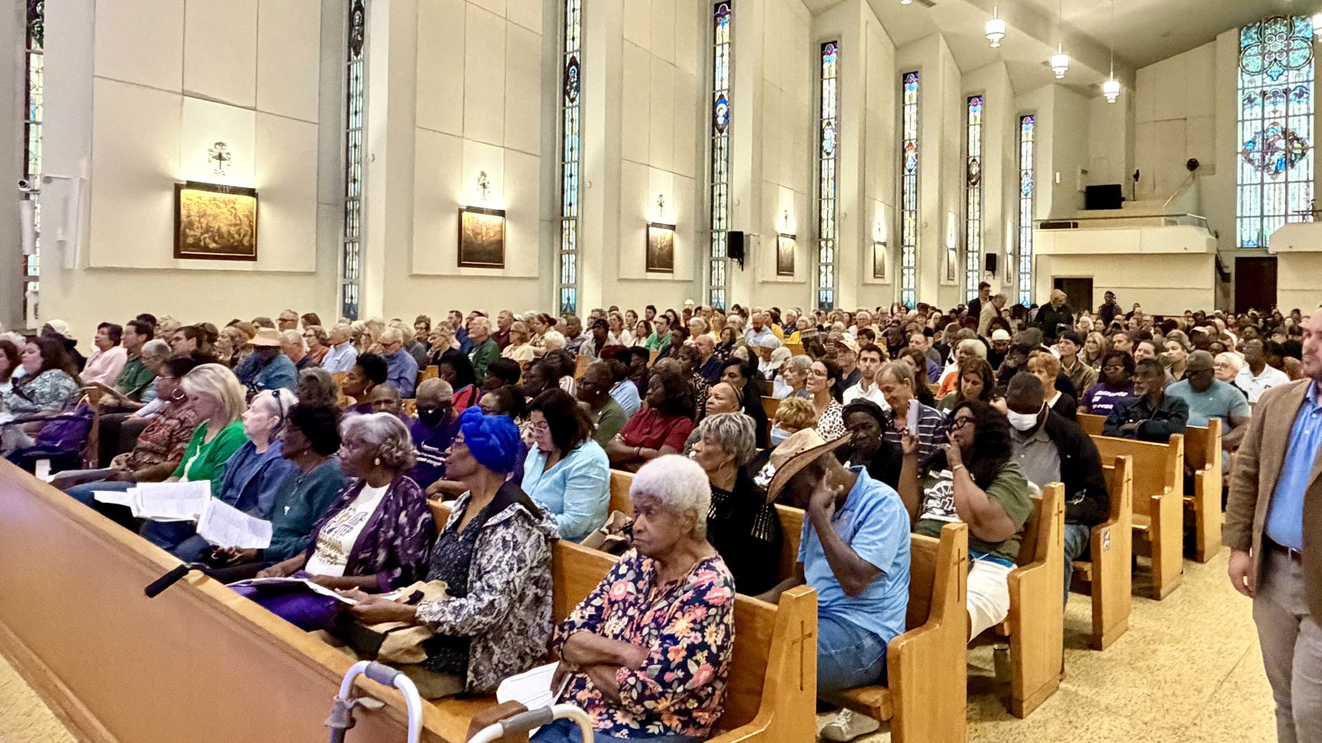 Inside a church filled with a diverse congregation seated in light wood pews, white walls, tall stained-glass windows, and hanging pendant lights.