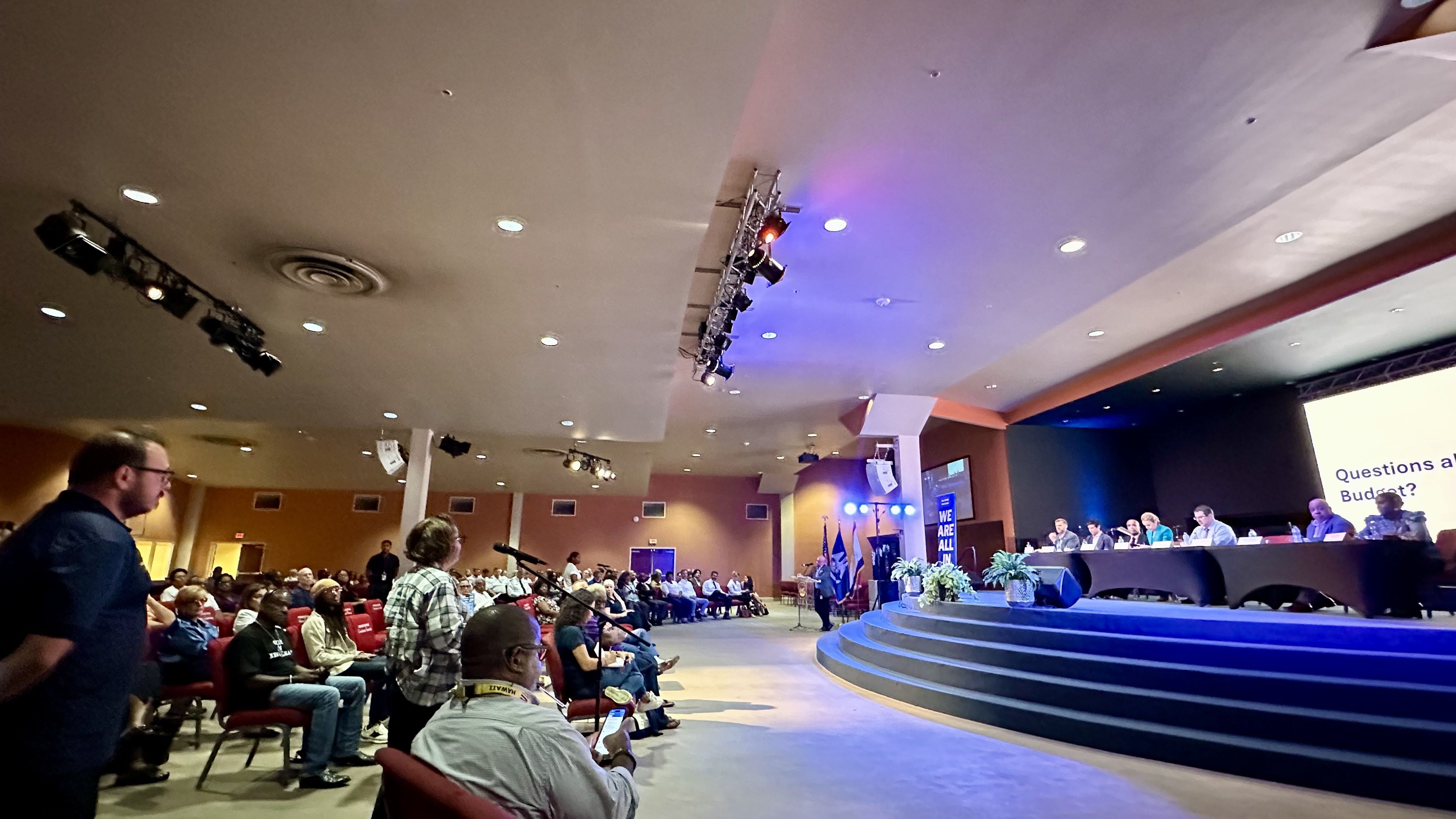 Audience in a large conference hall watches a panel of speakers on a curved blue-lit stage; overhead lighting rigs, ceiling lights, and attendees seated, some checking phones.
