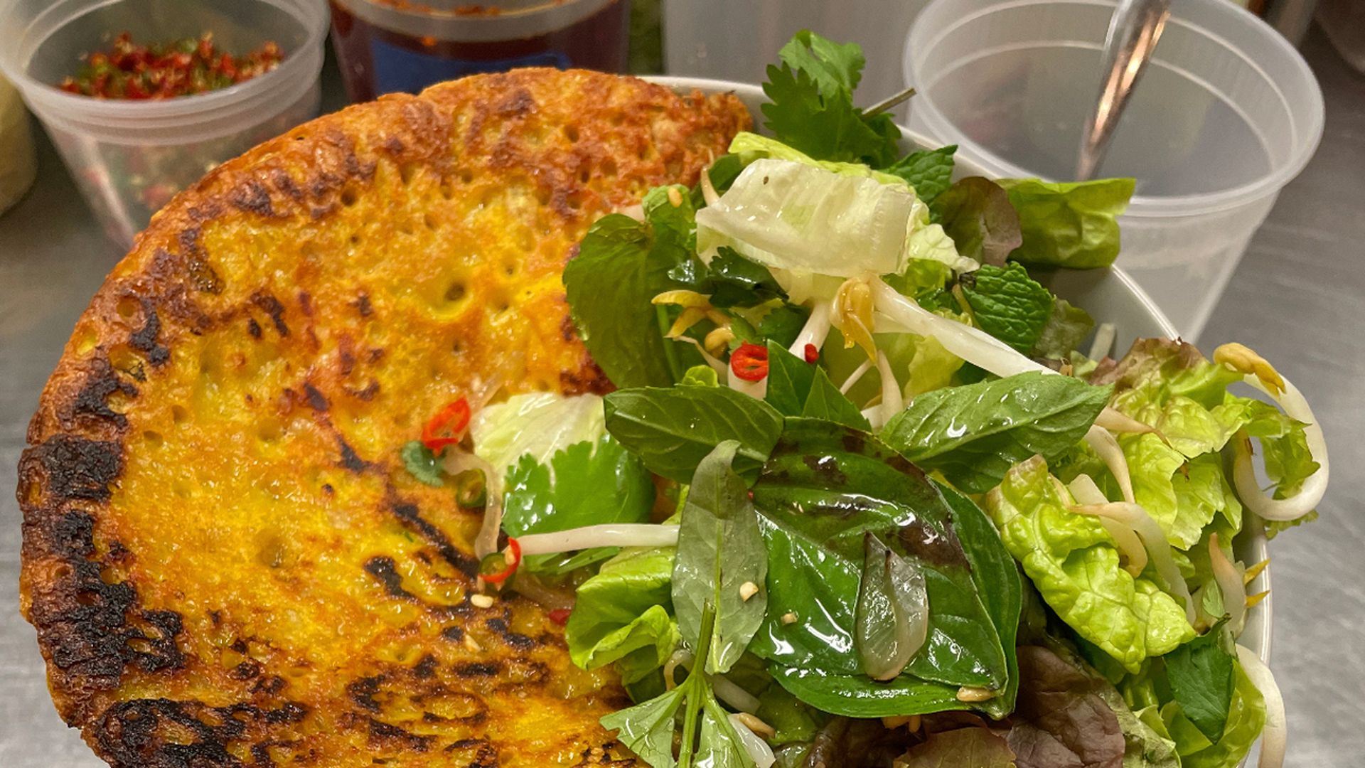 Close-up of a golden brown crispy rice cake with a side of fresh green leafy salad containing bean sprouts, mint, and chili slices, served in a white bowl on a metal surface.