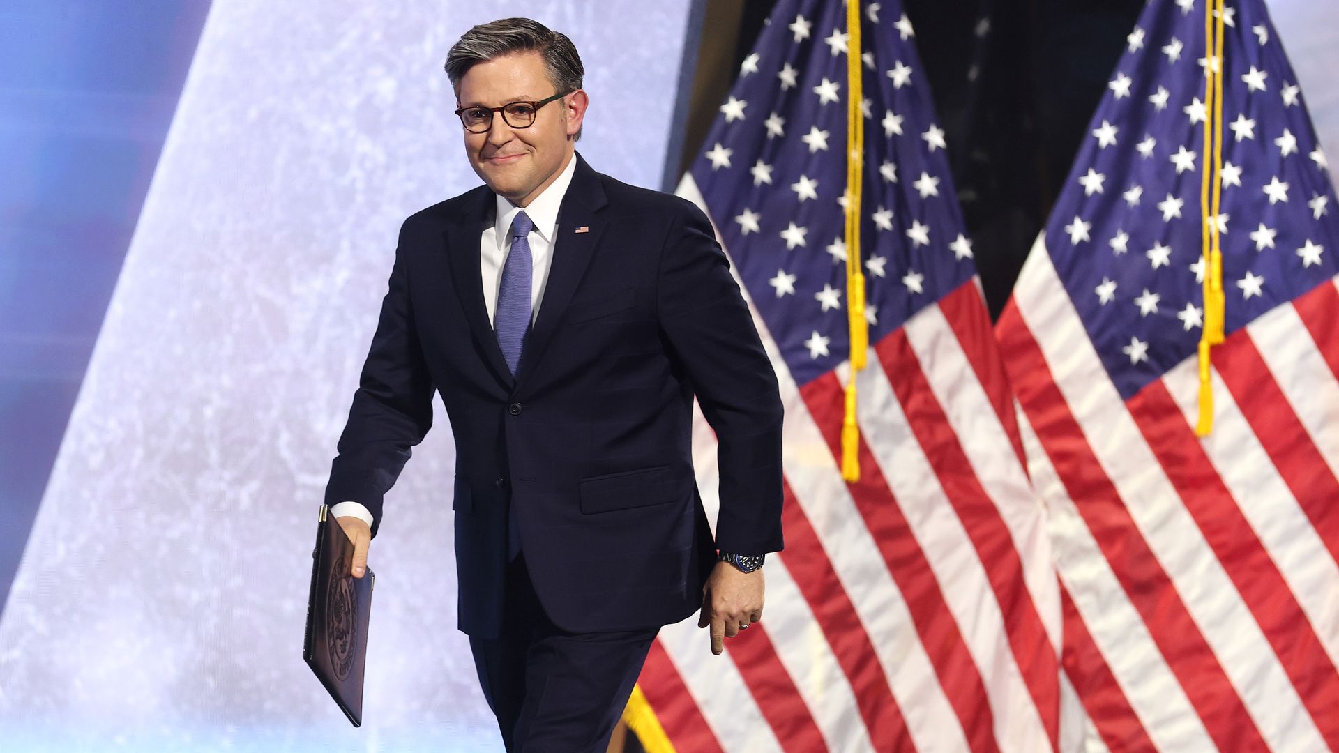 A man in a dark suit and glasses, wearing a purple tie, walking on stage while holding a portfolio; American flags line the background.