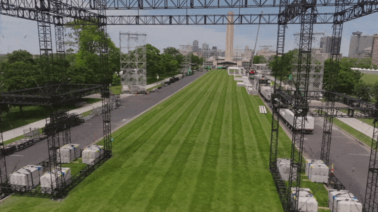 Outdoor event setup with black metal trusses framing a long, striped green lawn; trucks and equipment line the sides, with a city skyline and a tall monument visible in the distance.