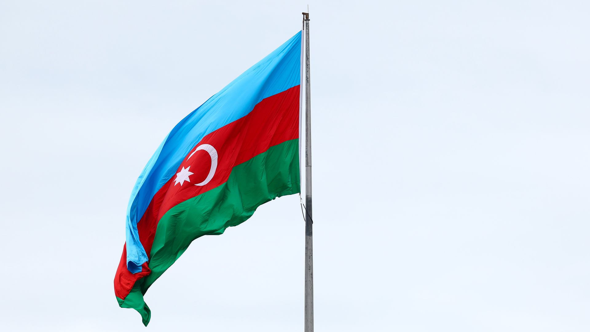 A flag with horizontal light blue, red, and green stripes, featuring a white crescent and an eight-pointed star in the center of the red stripe, flying on a metal pole against a cloudy sky.